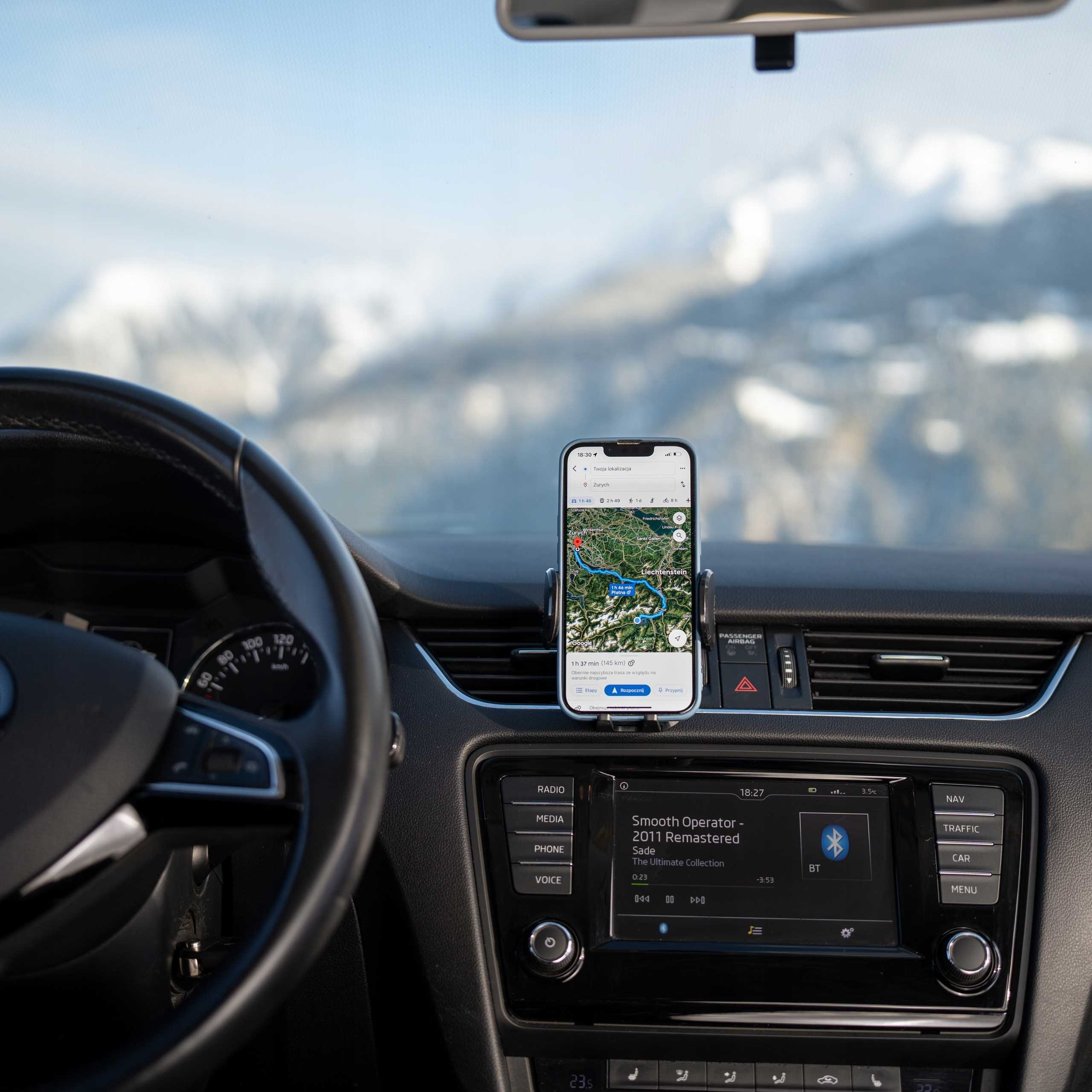 Wnętrze samochodu z telefonem z mapą nawigacji, górami w tle. Kierownica z przodu.
