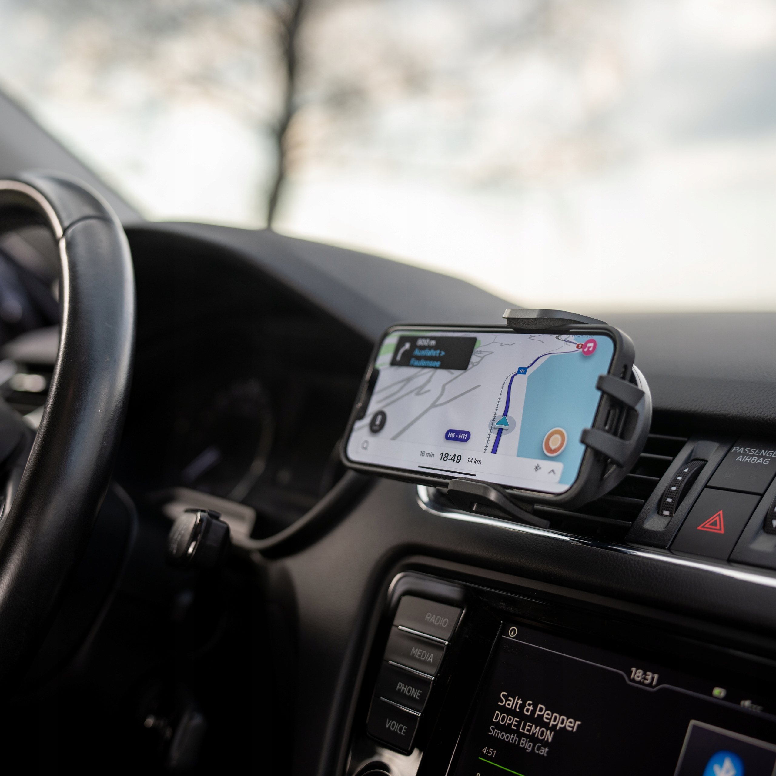 Wnętrze samochodu z telefonem pokazującym nawigację. Kierownica po lewej, deska rozdzielcza z ekranem i przyciskami. Zewnętrze jest rozmyte.