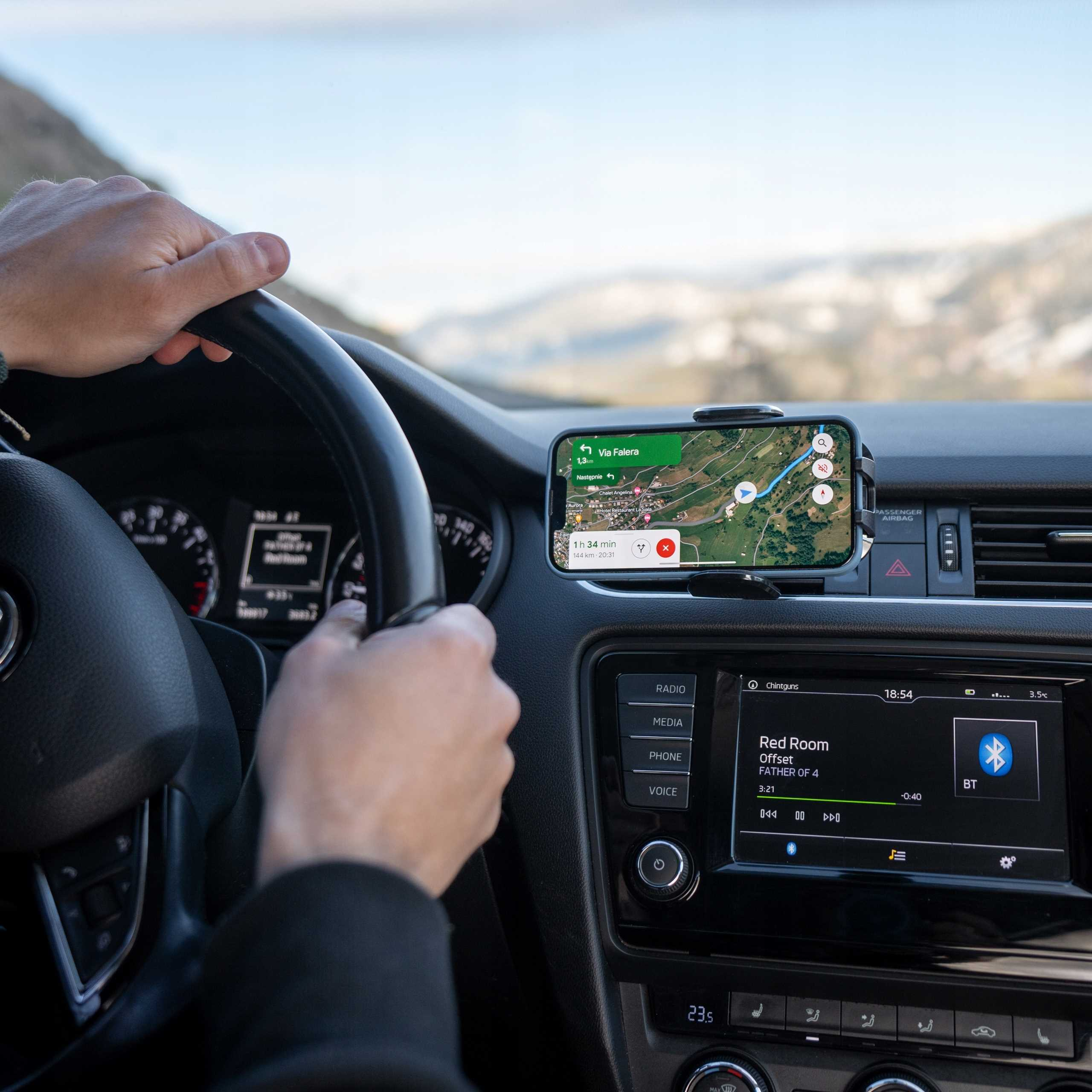 Ręce na kierownicy, telefon z mapą, deska rozdzielcza z radiem i muzyką, góry w tle, wnętrze samochodu.