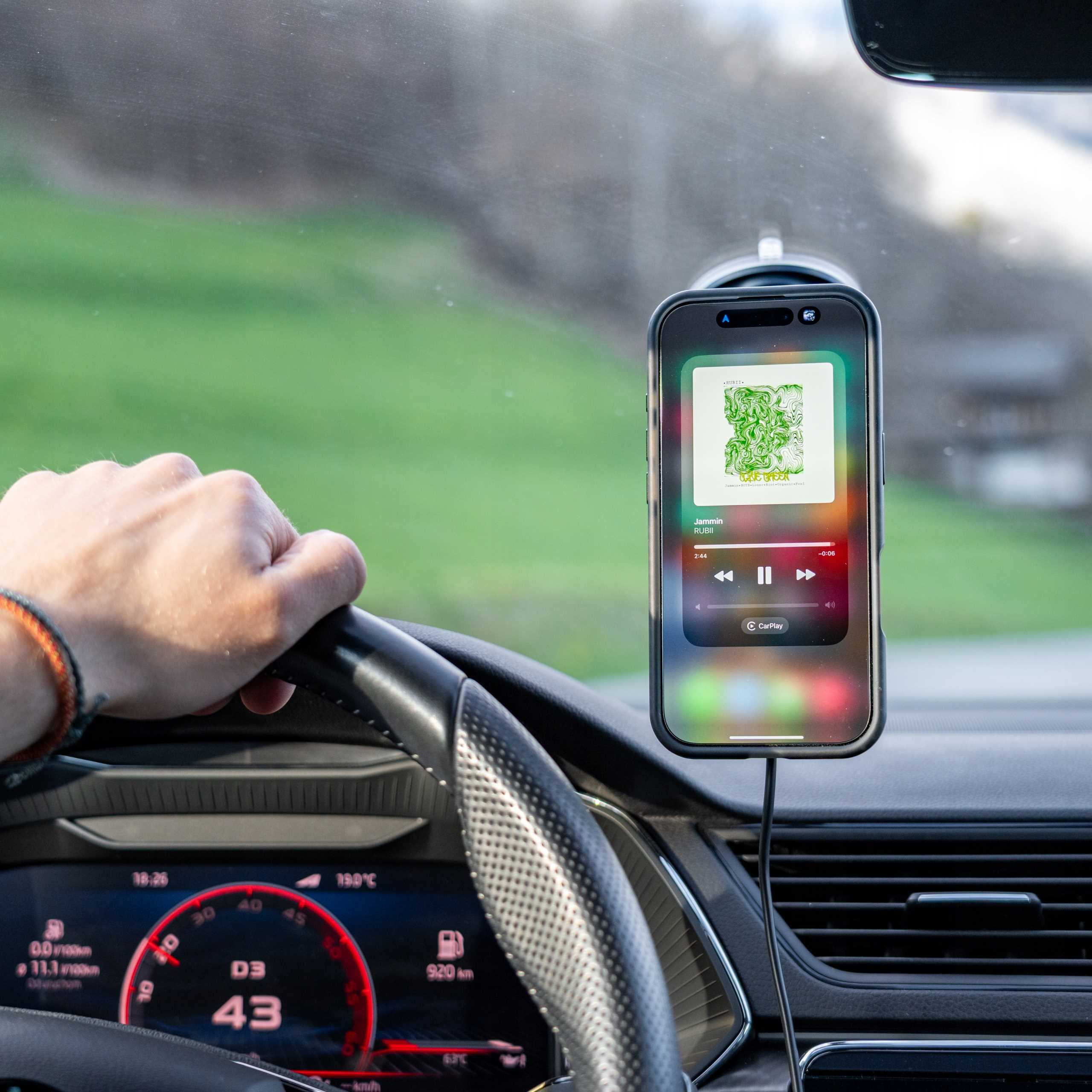 Osoba prowadzi samochód z telefonem wyświetlającym muzykę. Widoczna deska rozdzielcza i zielony krajobraz.