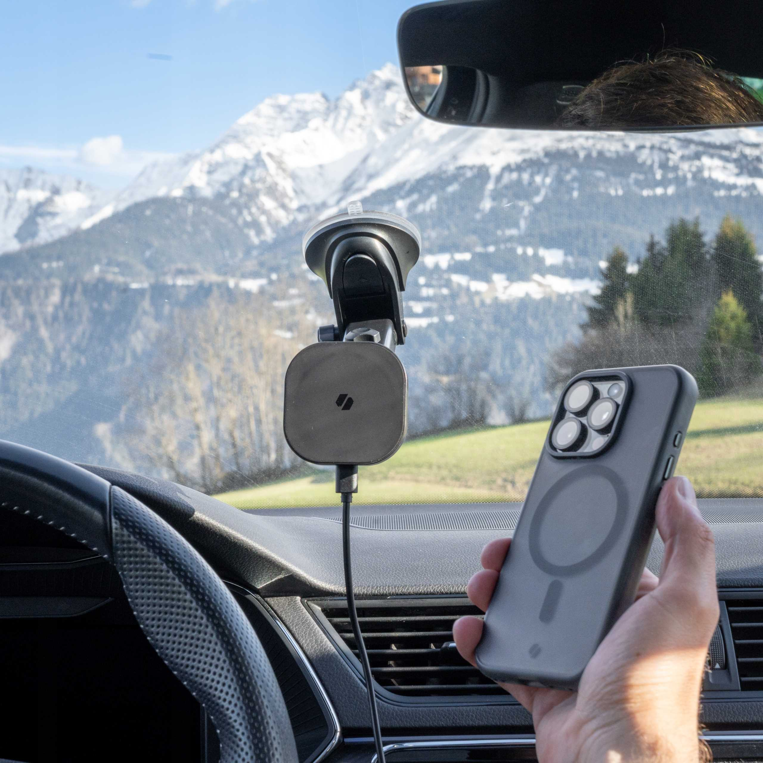 Ręka trzymająca telefon z etui w samochodzie. Uchwyt na telefon jest przymocowany do przedniej szyby. Na zewnątrz widać góry.