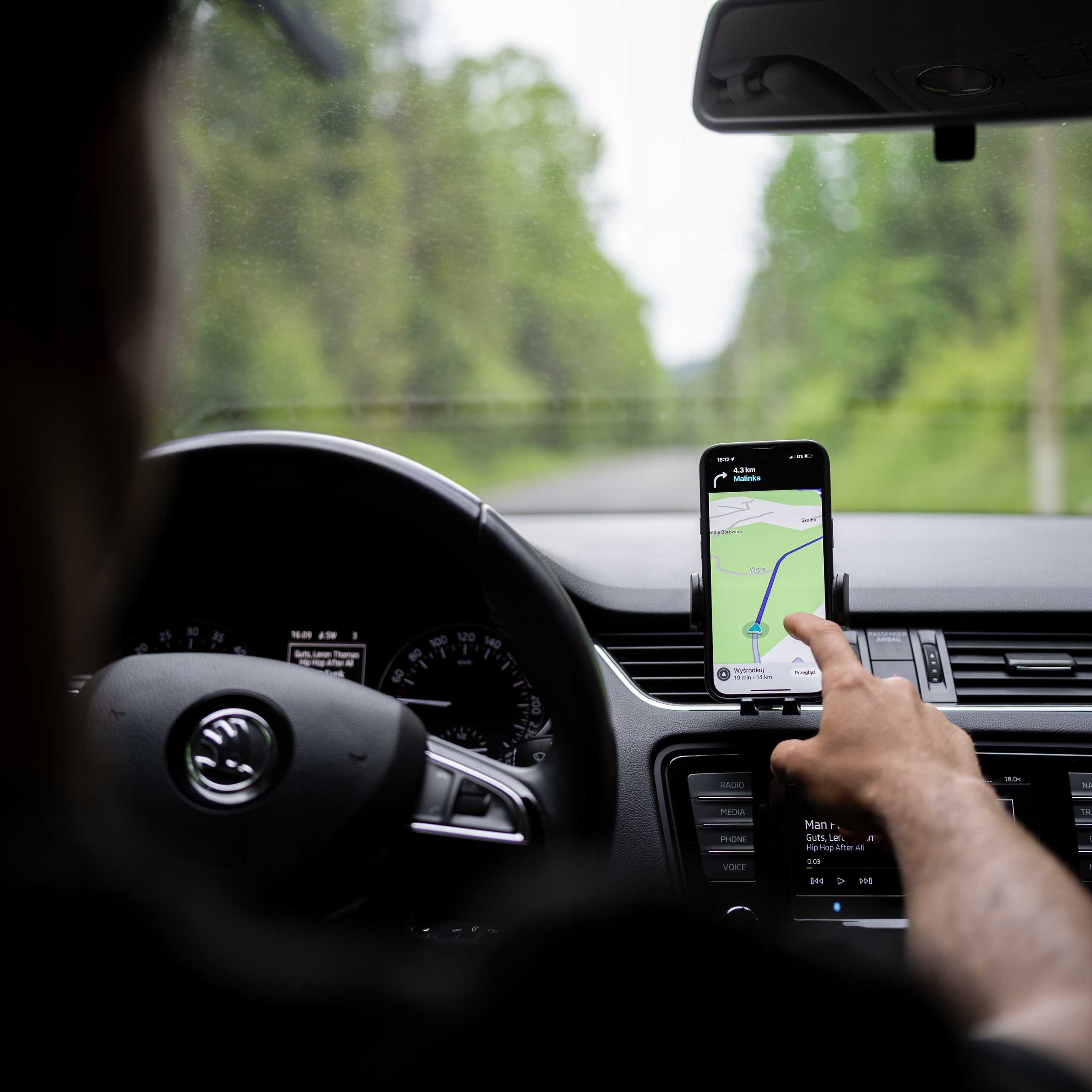 Osoba w samochodzie korzysta z telefonu z aplikacją nawigacyjną. Widoczna kierownica i deska rozdzielcza.