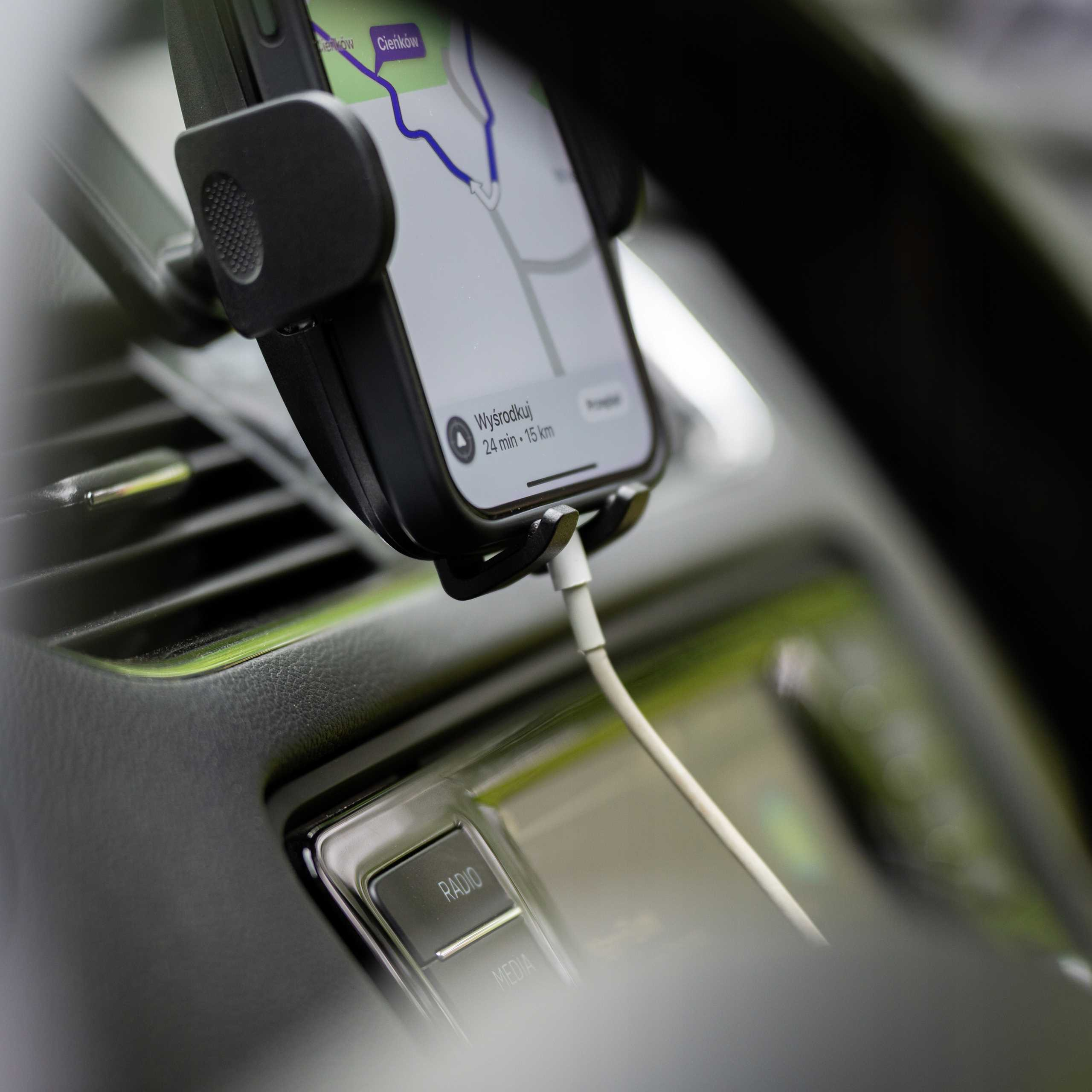 Telefon w uchwycie samochodowym wyświetla mapę. Podłączony jest kabel ładujący. Deska rozdzielcza ma przyciski radia i multimediów.