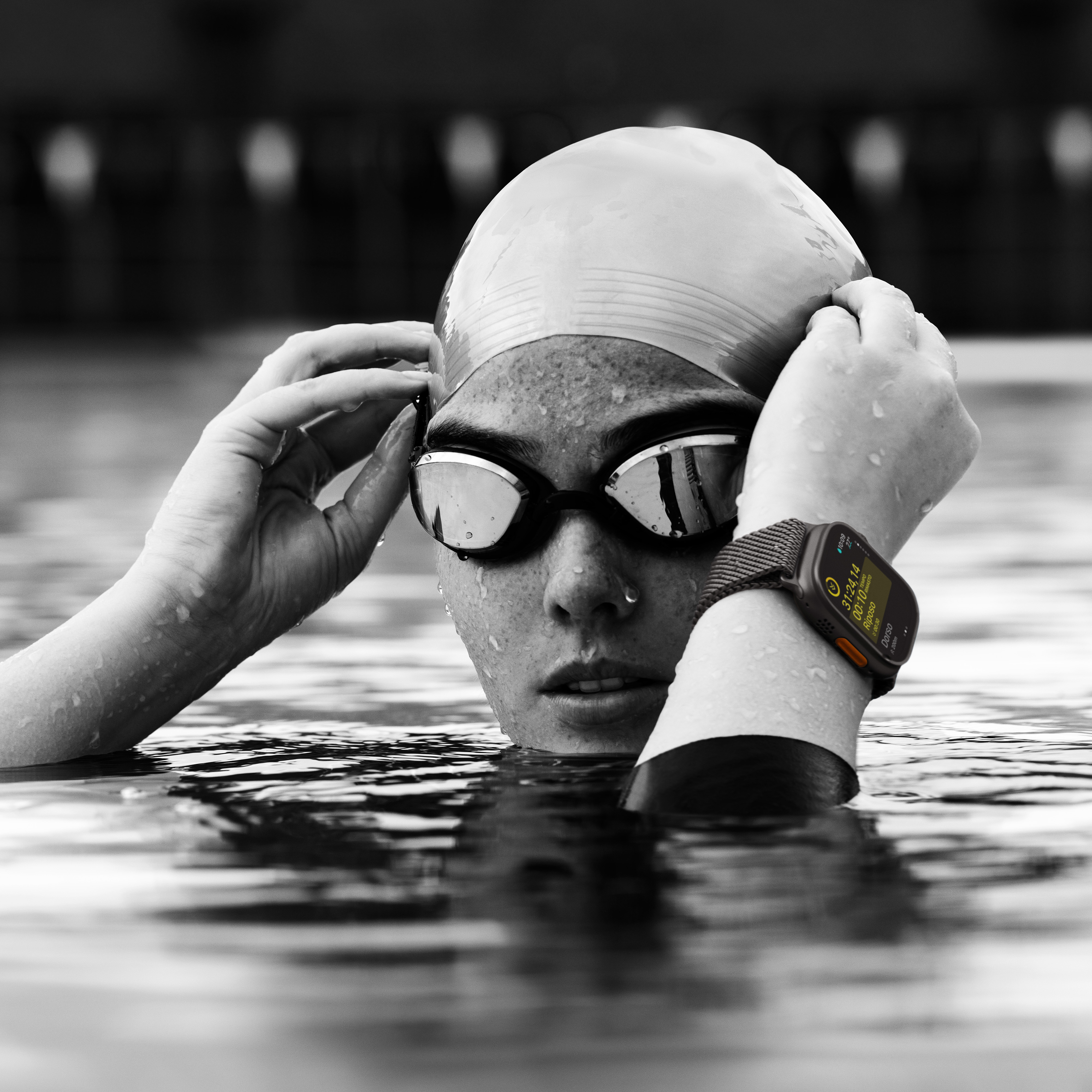 Pływak wyłania się z wody, w okularach i czepku. Widoczny jest smartwatch. Obraz monochromatyczny.