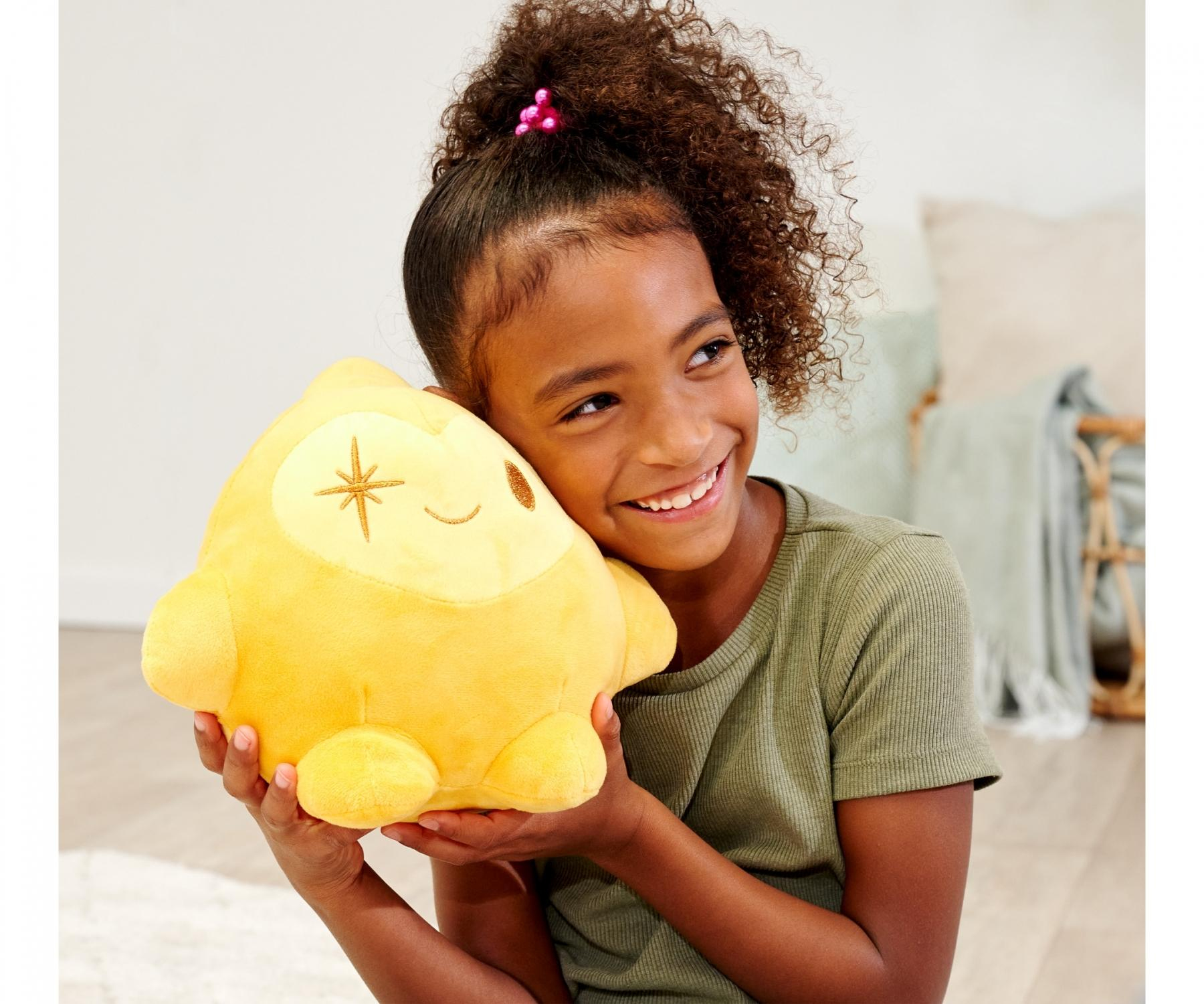Uśmiechnięta dziewczyna trzyma żółtą pluszową zabawkę. Zabawka ma gwiazdę i twarz. Dziewczyna ma na sobie zieloną koszulę, a w tle jest pokój.