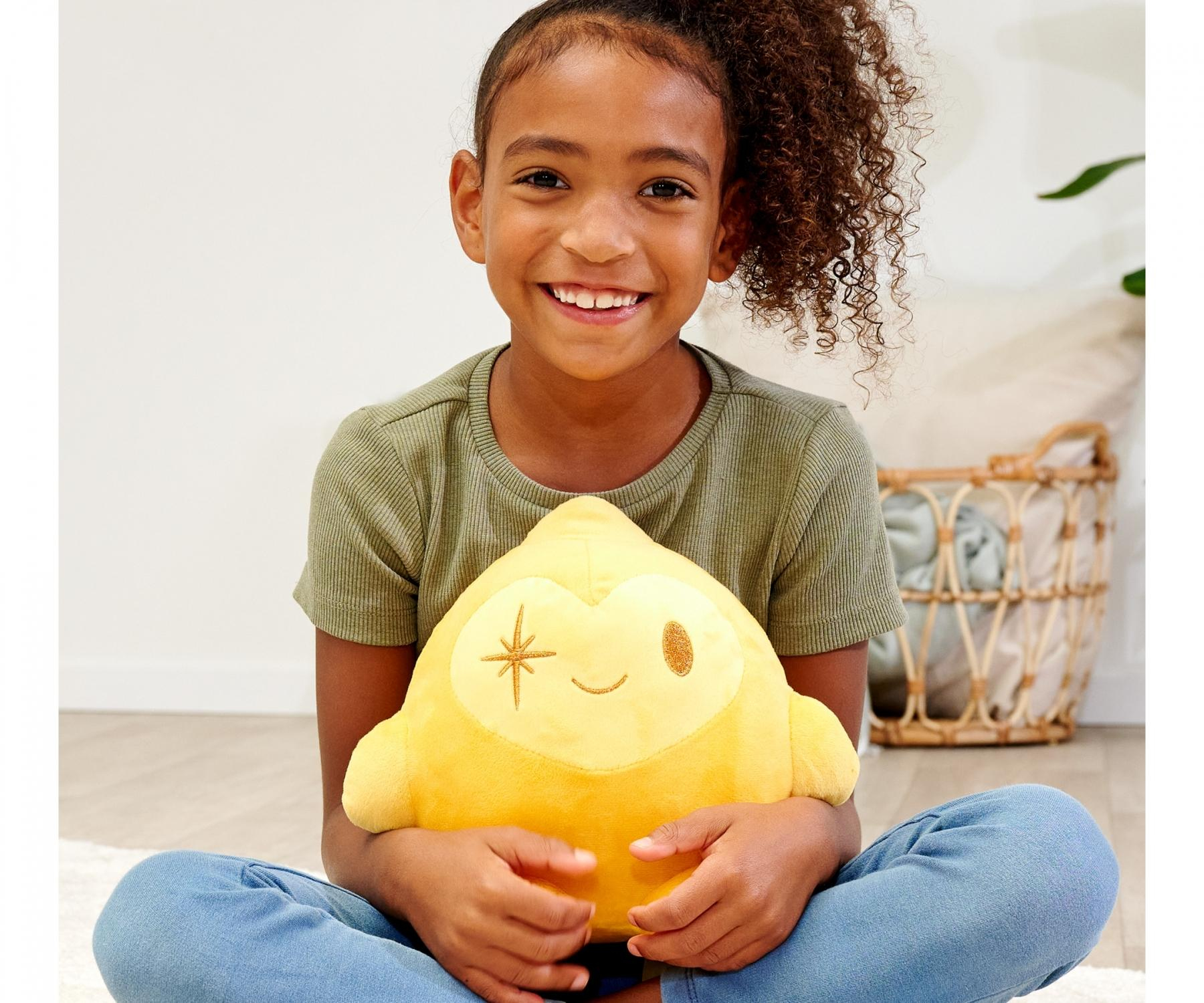 Uśmiechnięta dziewczyna siedzi ze skrzyżowanymi nogami, trzymając żółtą pluszową zabawkę. Ma na sobie zieloną koszulę i niebieskie dżinsy, w tle kosz.