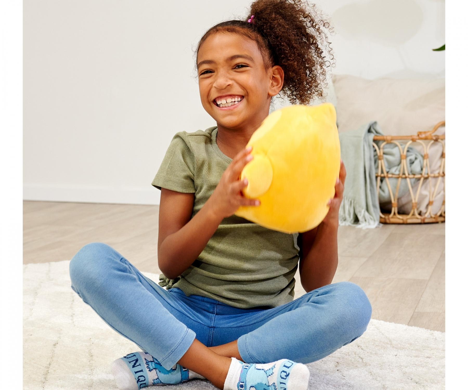 Uśmiechnięta dziewczynka siedzi ze skrzyżowanymi nogami, trzymając żółtą pluszową zabawkę. Ma na sobie niebieskie dżinsy i zieloną koszulę, na białym dywanie.