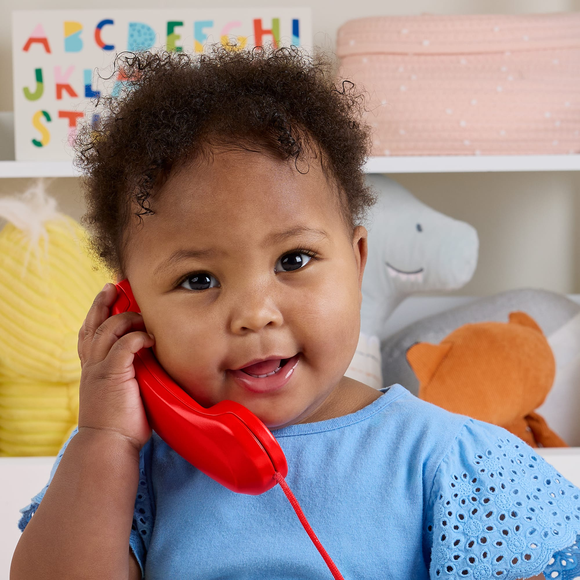 Ein lächelndes Baby hält ein rotes Telefon an sein Ohr. Im Hintergrund sind eine farbenfrohe Alphabet-Tafel und Spielzeug.