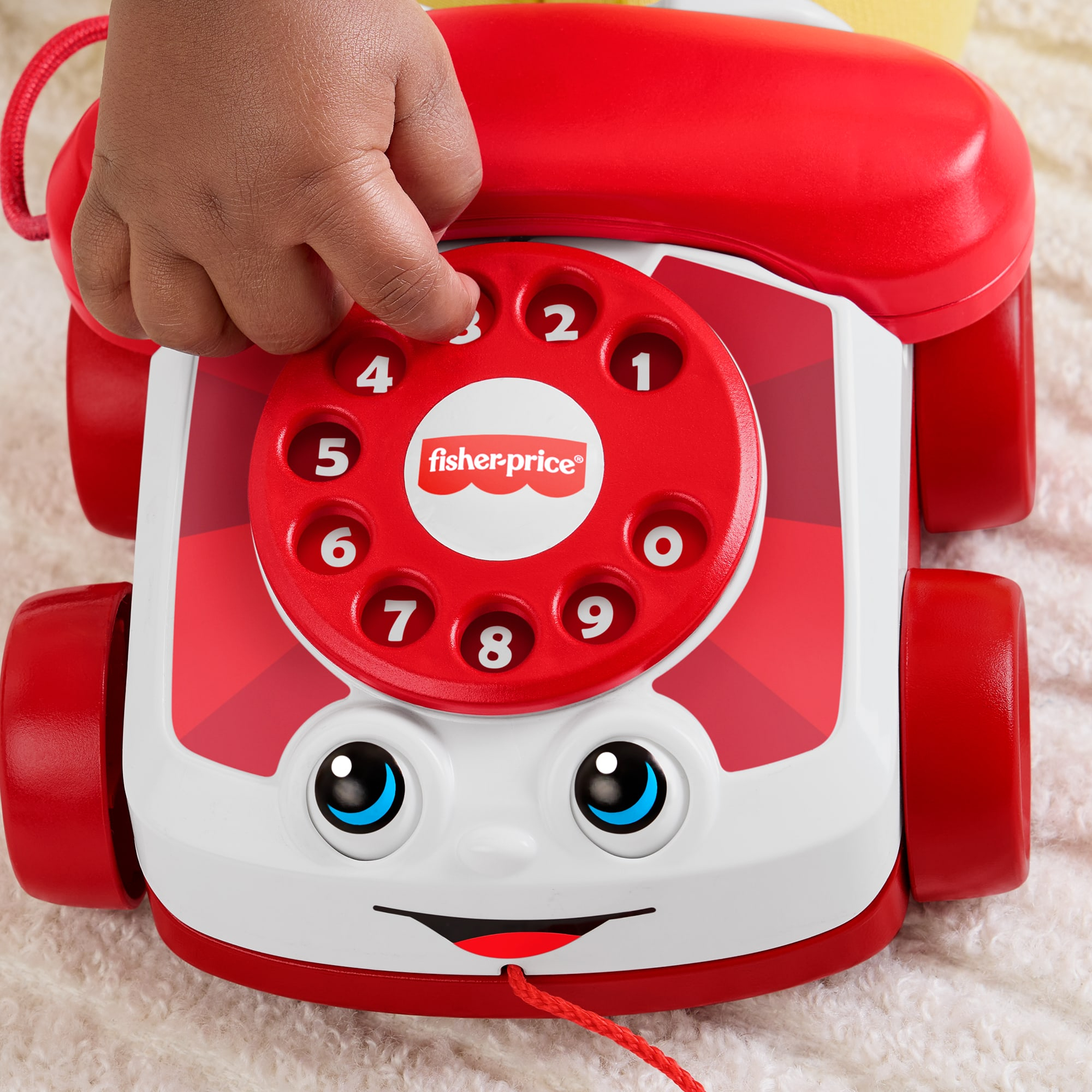 Die Hand eines Kindes drückt die Taste '3' auf einem rot-weißen Spielzeugtelefon von Fisher-Price. Das Telefon hat ein lächelndes Gesicht und Räder.