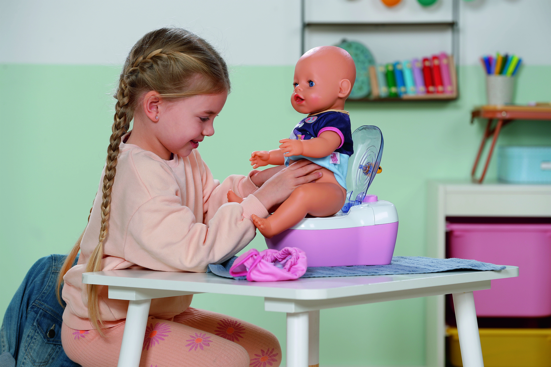 Dziewczyna z warkoczami pomaga lalce na nocniku. Lalka ma na sobie niebieską koszulę. Scena rozgrywa się w pokoju z zabawkami.