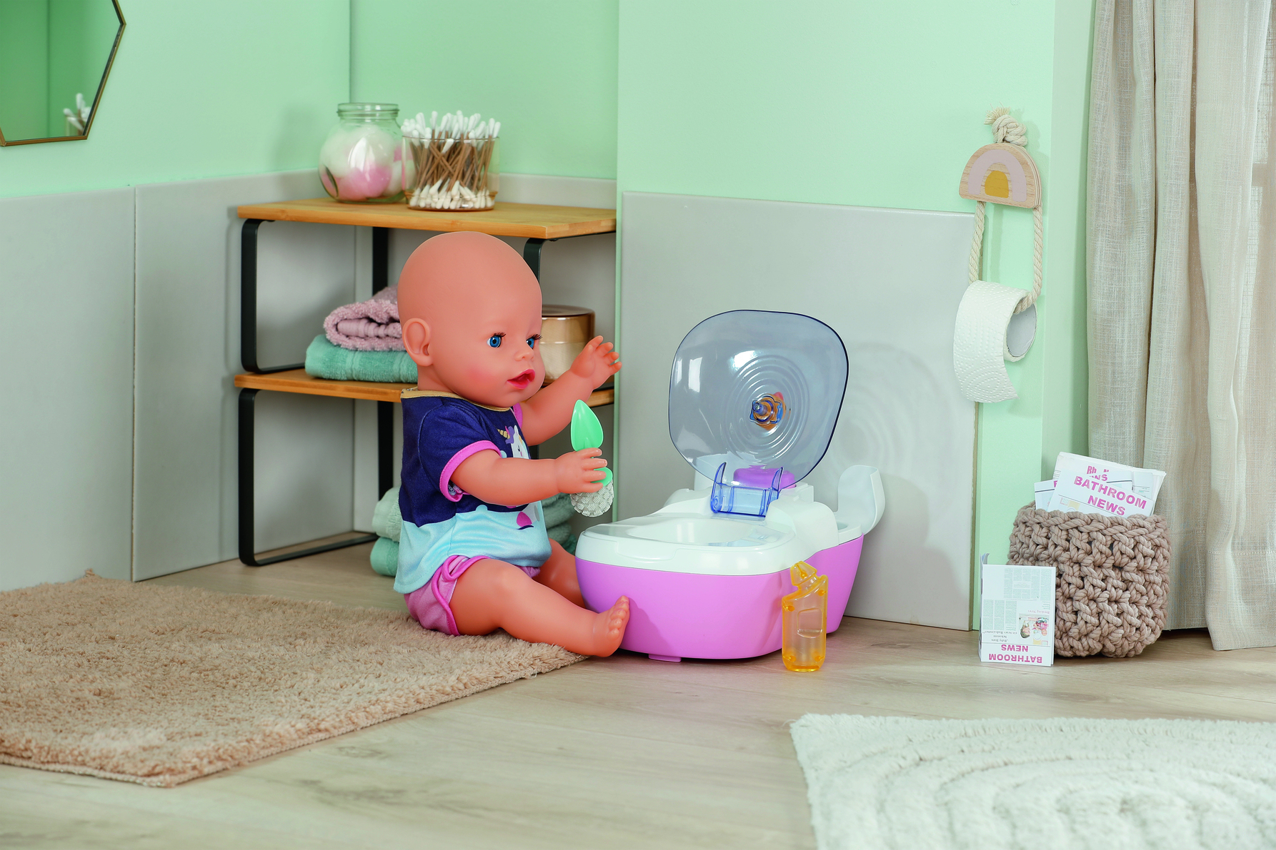 Lalka siedzi obok różowego nocnika z przezroczystą pokrywką. W pobliżu znajduje się mała butelka i zielona zabawka. Scena to łazienka.