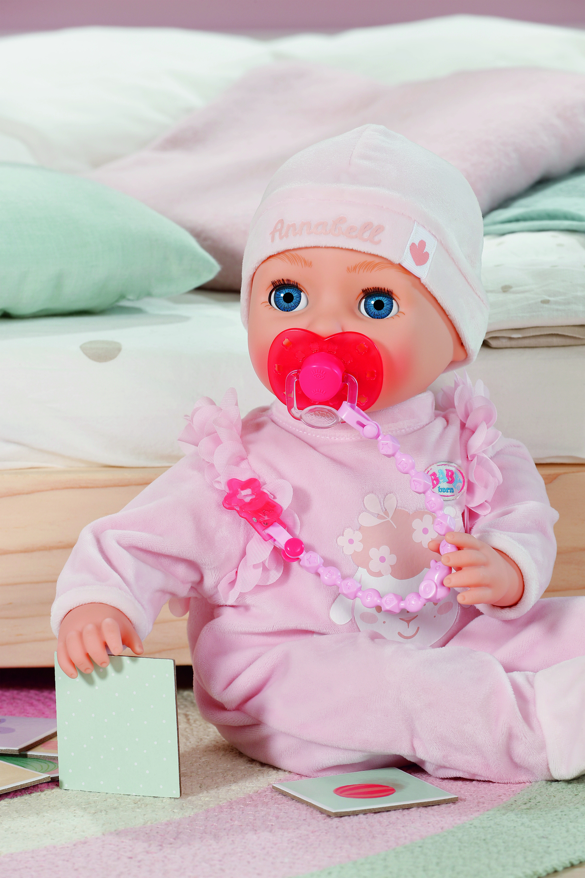 Eine Babypuppe in rosa Kleidung mit Hut und Schnuller. Sie sitzt auf einem gestreiften Teppich, mit Karten und Kissen im Hintergrund.