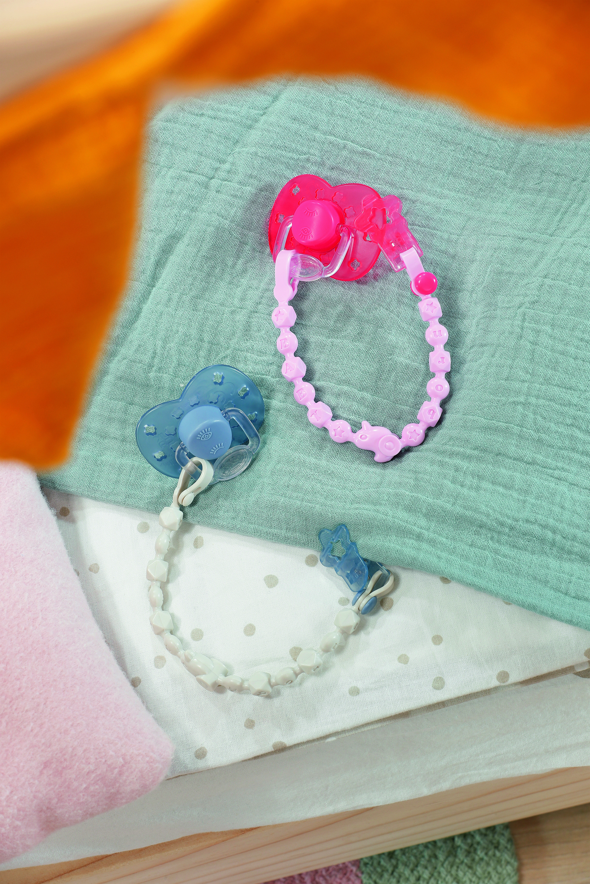 Zwei Schnuller mit Ketten, einer rosa und einer blauen, auf einem grünen Tuch, mit einem orangefarbenen Tuch in der oberen linken Ecke.