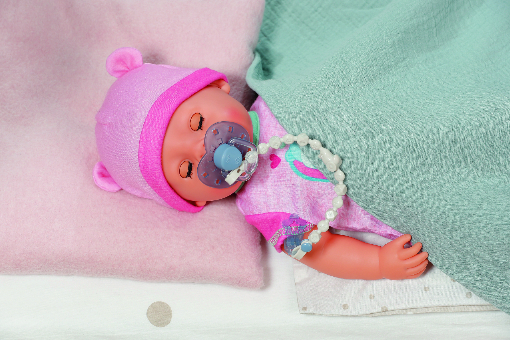 Eine Babypuppe mit rosa Mütze und Kleidung, Schnuller und Perlenkette, liegt auf einem rosa Kissen und einer hellgrünen Decke.