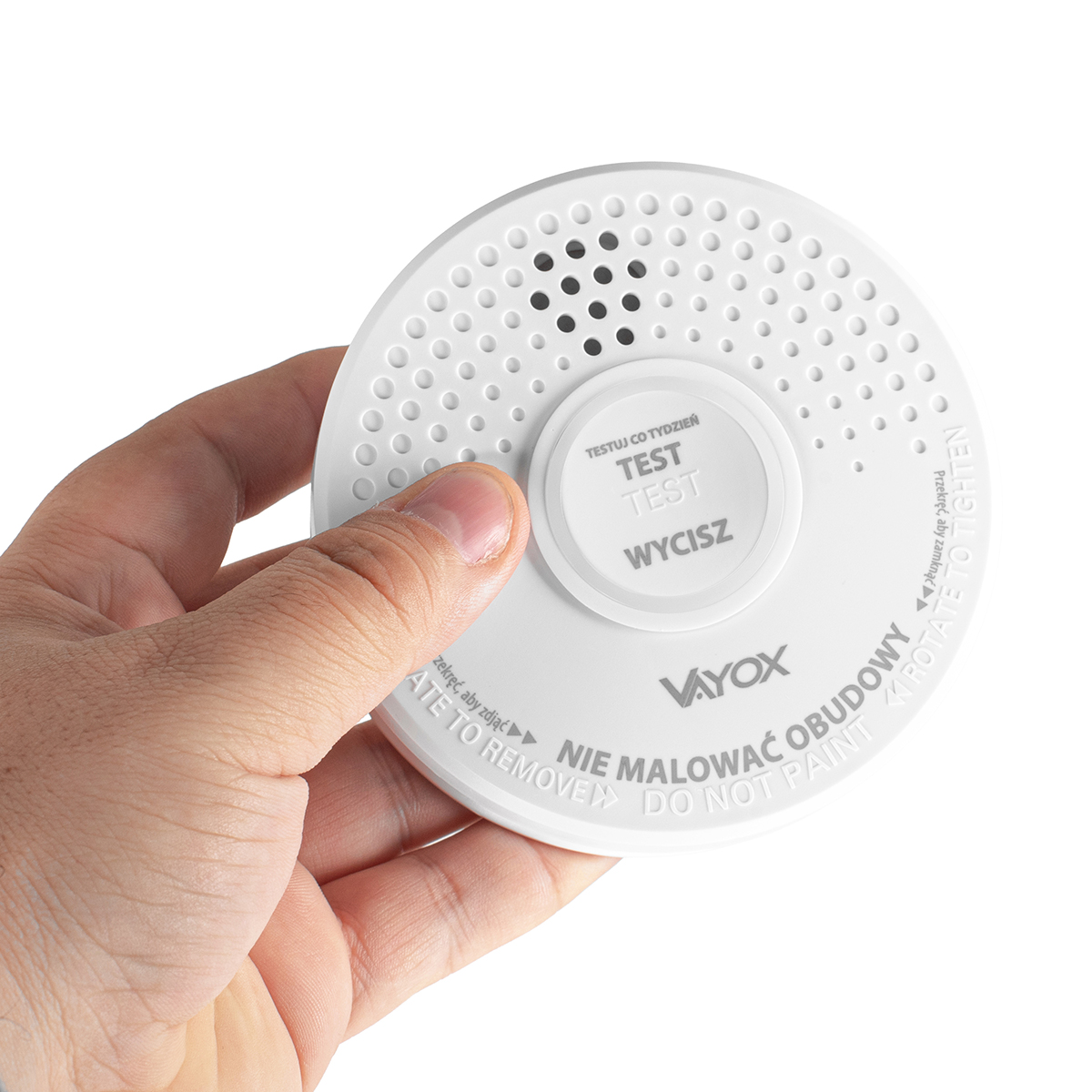 Ręka trzyma białą czujkę dymu. Urządzenie ma przycisk testu i tekst w języku polskim. Tło jest białe.