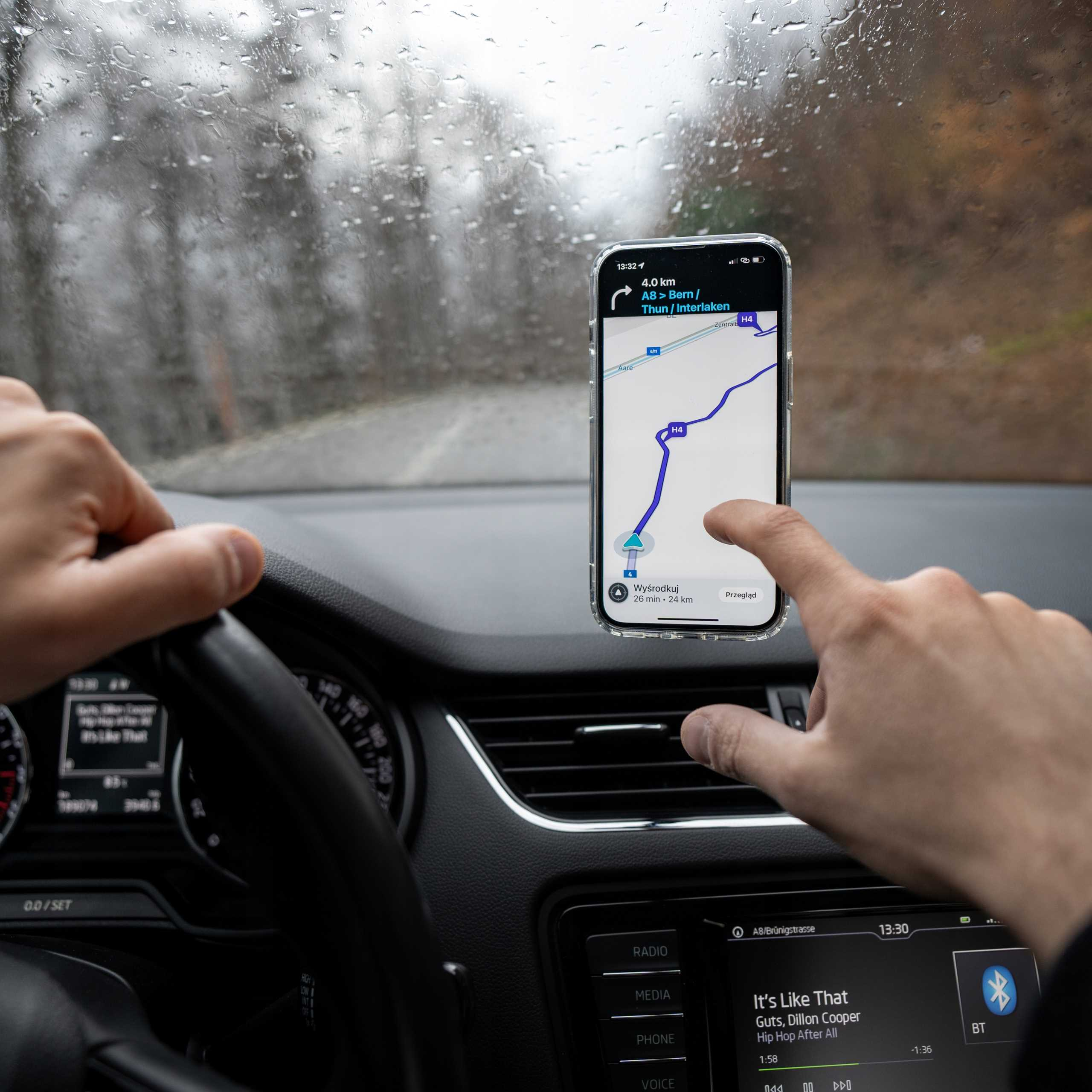 Osoba używa telefonu z nawigacją w samochodzie. Ekran pokazuje mapę. Krople deszczu na przedniej szybie.