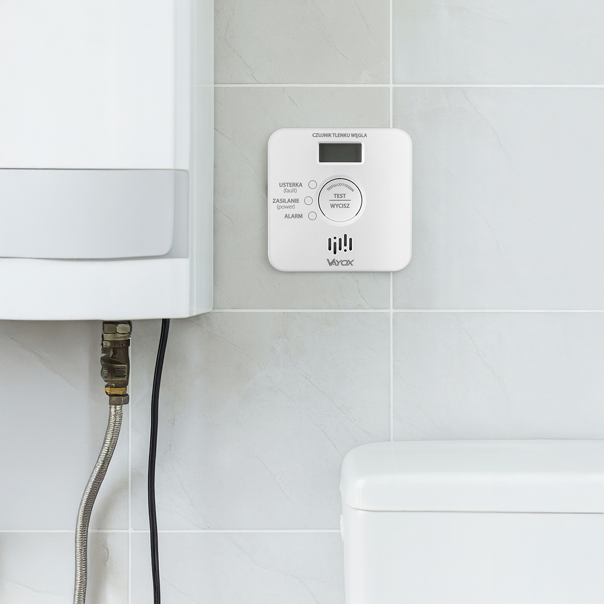 Biały czujnik tlenku węgla jest zamontowany na kafelkowej ścianie w pobliżu białego podgrzewacza wody i toalety.