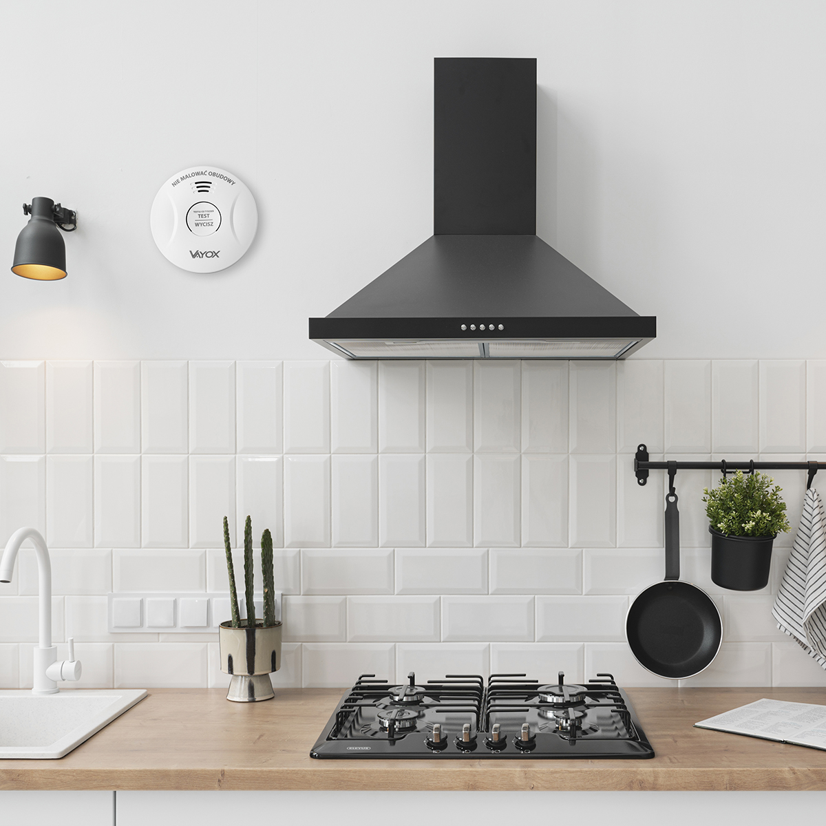Scena kuchenna z białymi kafelkami, czarną kuchenką i czujnikiem dymu. Nad kuchenką znajduje się czarny okap. Wiszą mała roślina i patelnia.