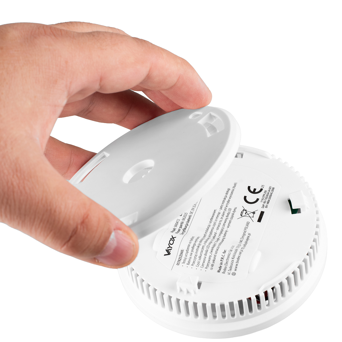 Ręka otwiera biały czujnik dymu. Ma okrągły kształt z otworami wentylacyjnymi i etykietę z tekstem oraz znakiem CE.