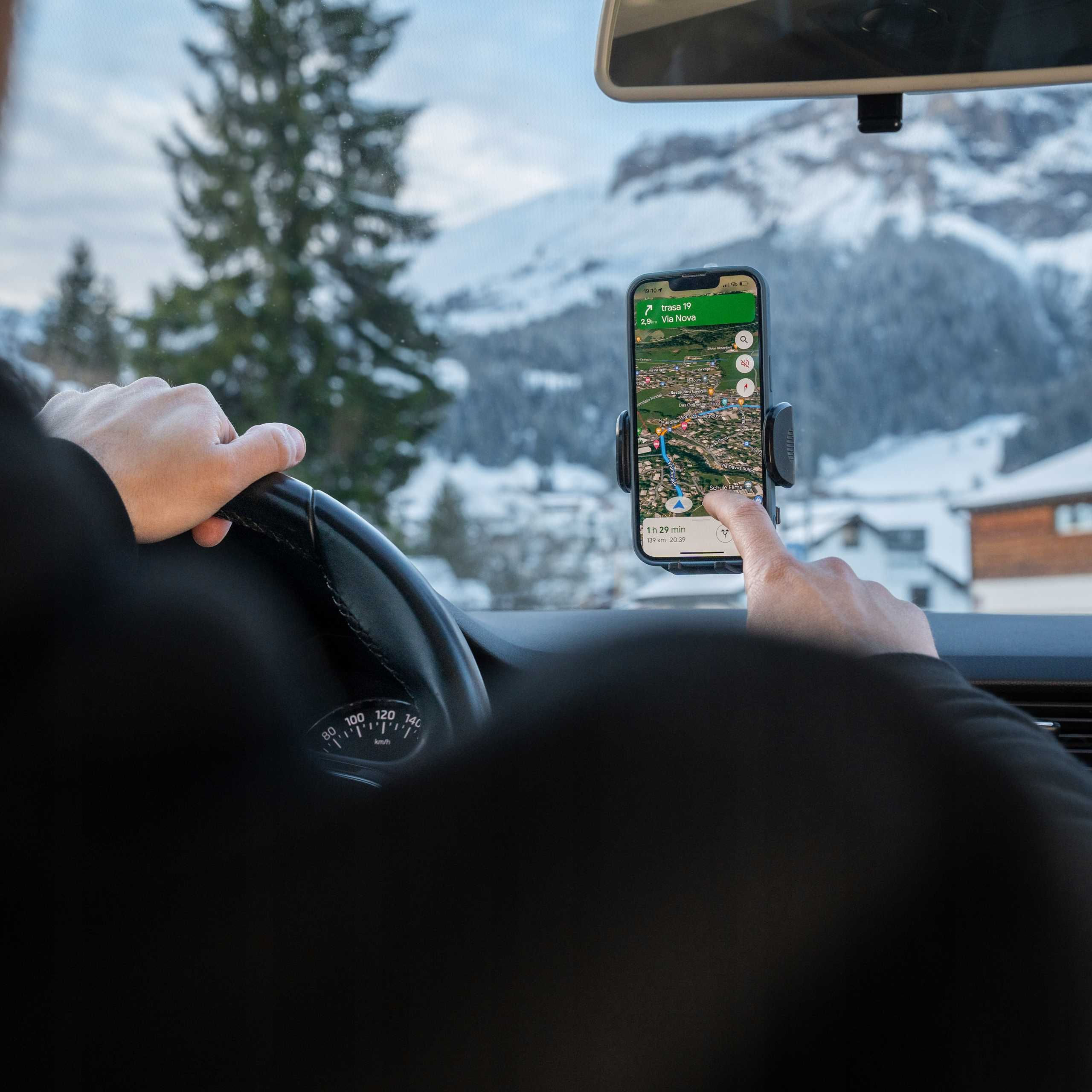 Osoba w samochodzie korzysta z GPS w telefonie. Tło przedstawia ośnieżone góry i drzewa.