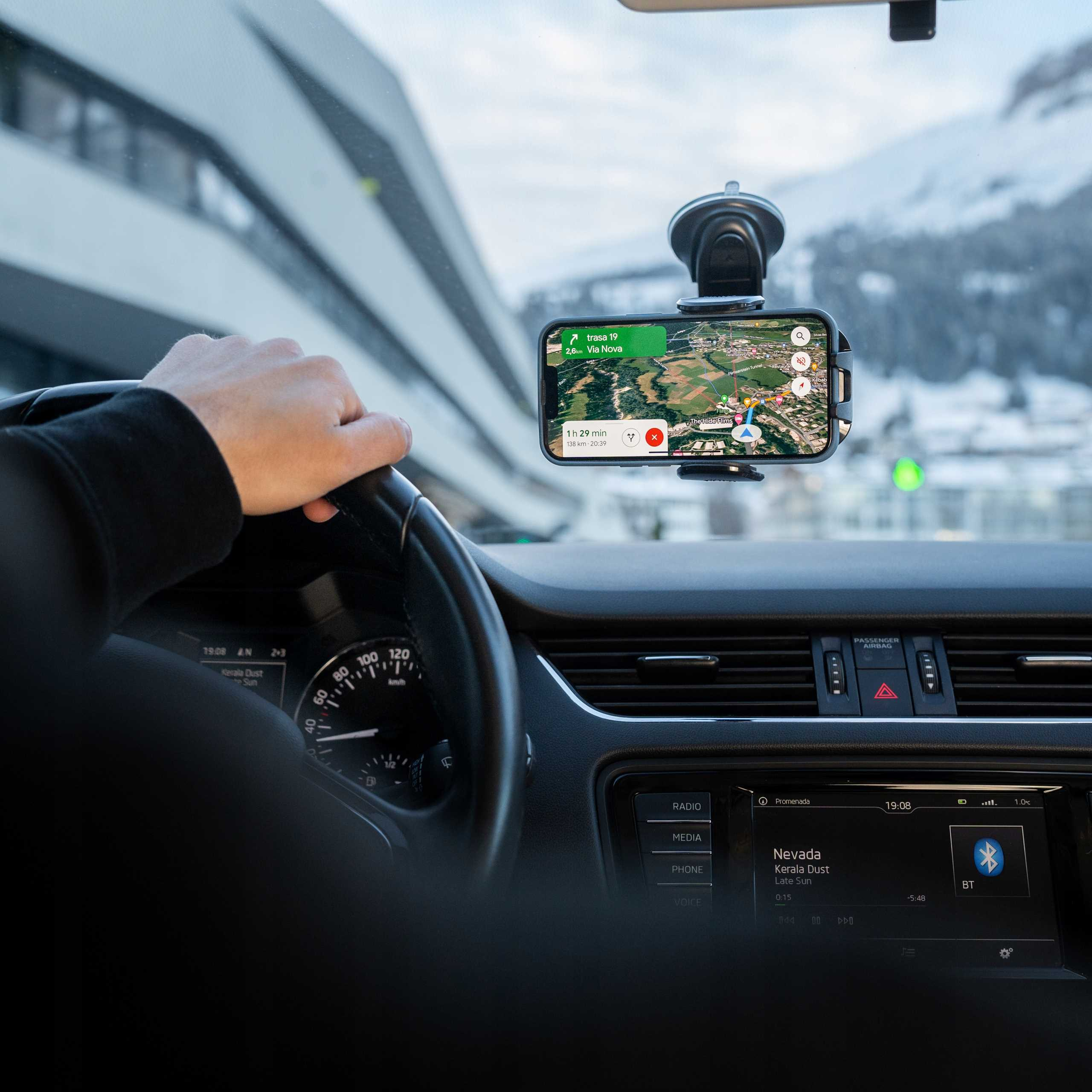 Osoba prowadzi samochód, korzystając z telefonu z nawigacją. Widoczna deska rozdzielcza i góry przez przednią szybę.