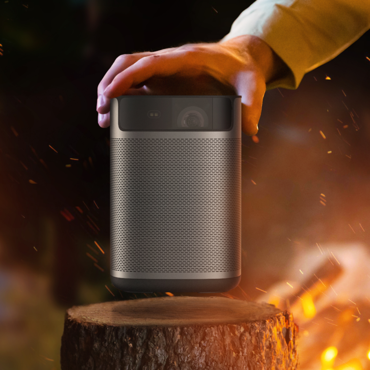 A hand holds a cylindrical speaker on a tree stump. Sparks and a campfire are in the background.