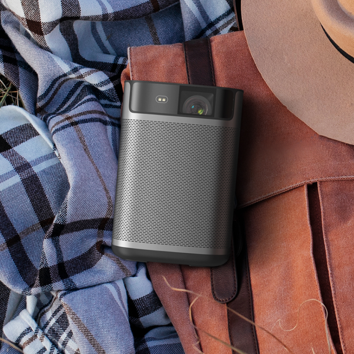 A silver projector rests on a brown bag, with a plaid blanket and a hat nearby. The projector is the central focus of the image.