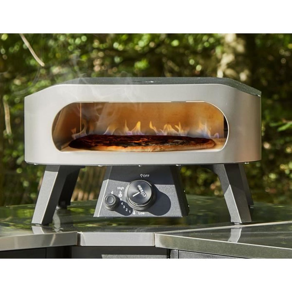 Un forno per pizza con pizza all'interno, fiamme visibili. Il forno è metallico, su un tavolo, con uno sfondo verde foglioso.