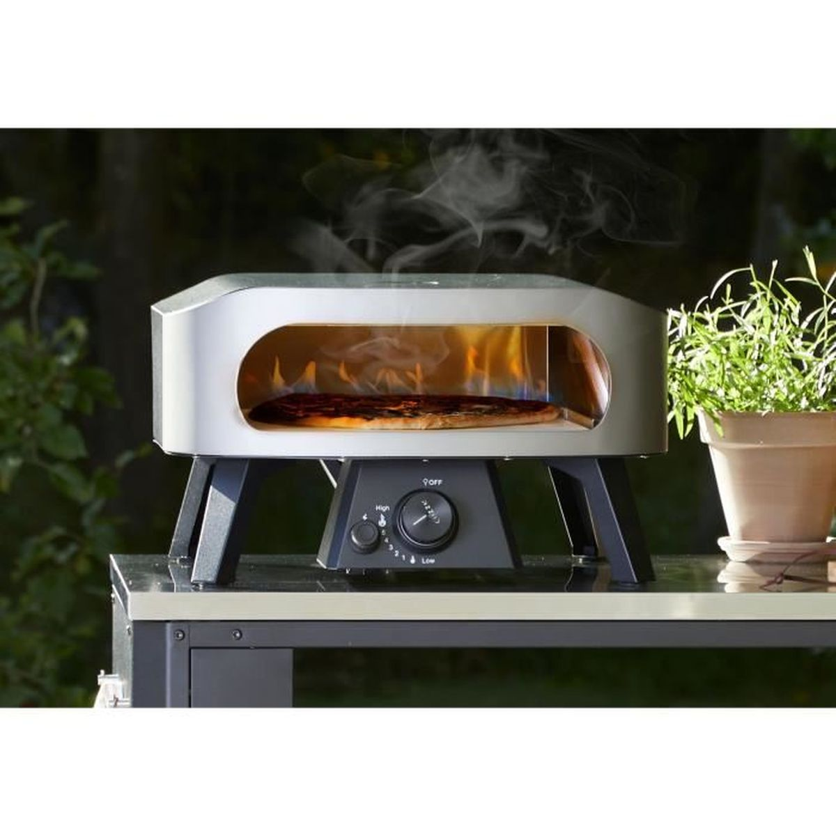 Un forno per pizza con una pizza all'interno e fiamme. Il forno è su un tavolo con un vaso di fiori a destra. Il fumo sale dal forno.