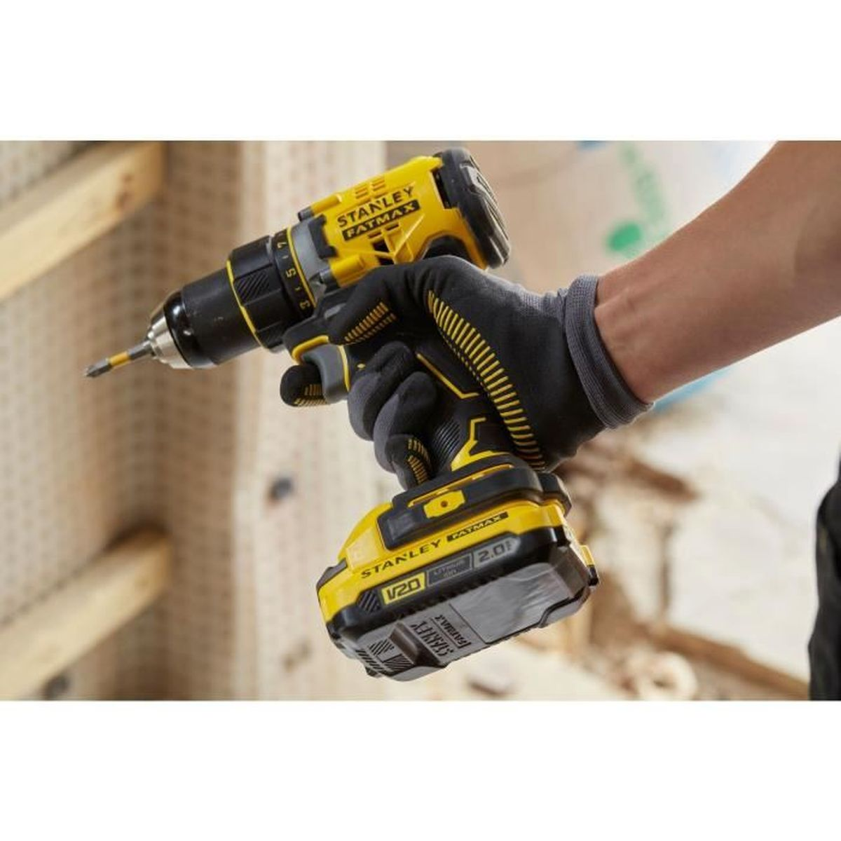 Una mano enguantada sostiene un taladro Stanley Fatmax amarillo y negro. El fondo incluye tablas de madera.