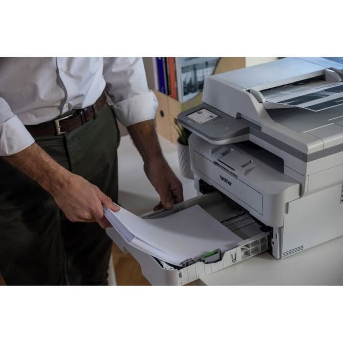 Mężczyzna w białej koszuli wkłada papier do drukarki. Drukarka jest szara i biała. W tle książki i roślina.