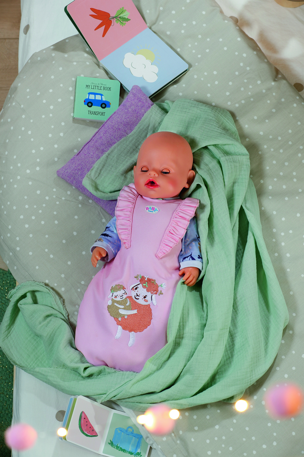 Lalka w różowym śpiworku z haftowaną owcą spoczywa na łóżku. Otaczają ją książki i zielony koc.
