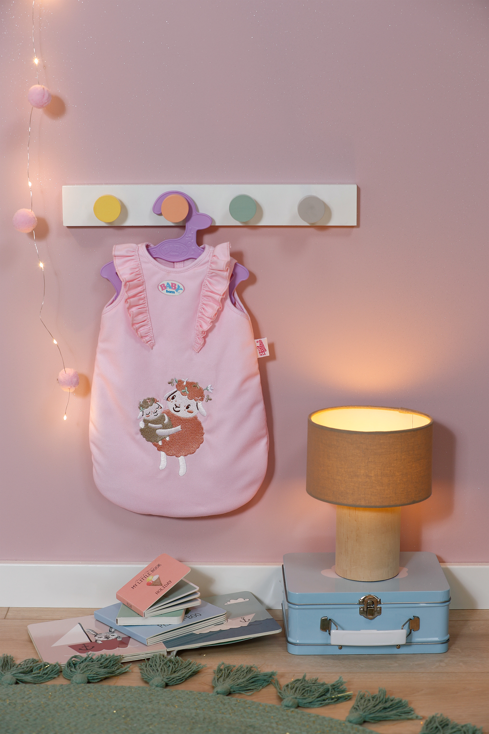 Różowy śpiwór dla dziecka wisi na ścianie. Lampa i książki leżą na podłodze. Ściana ma girlandę i półkę z kołkami.