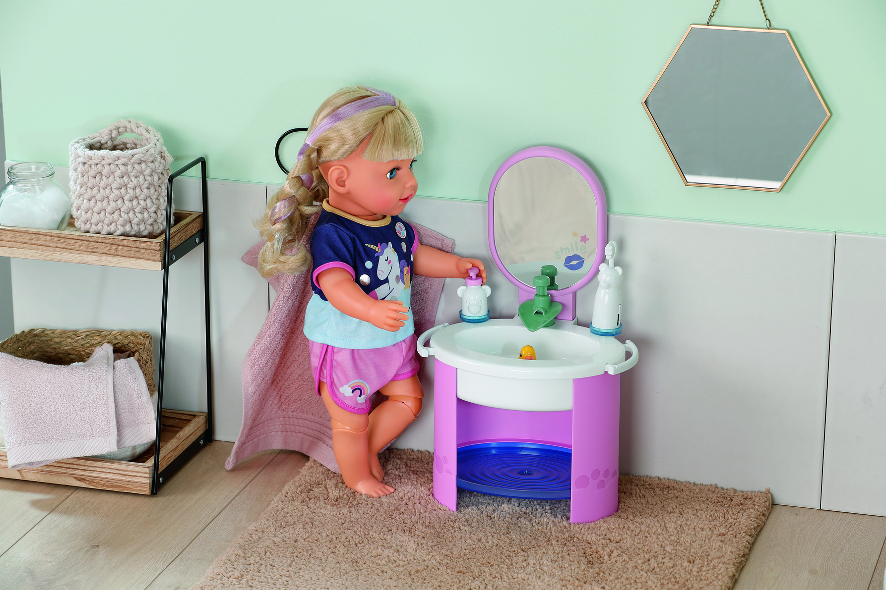 Lalka stoi w pobliżu zabawkowego zlewu. Scena obejmuje lustro, półkę z przedmiotami i dywan. Lalka ma blond włosy i nosi koszulę.