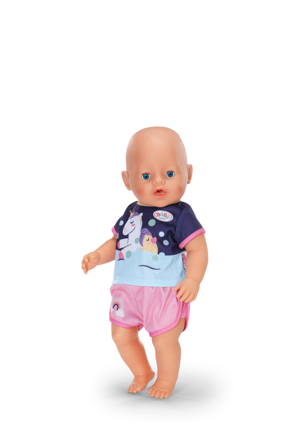Lalka stoi. Ma łysą głowę, niebieskie oczy i ma na sobie niebieską koszulę i różowe spodenki. Koszulka ma wzór jednorożca.