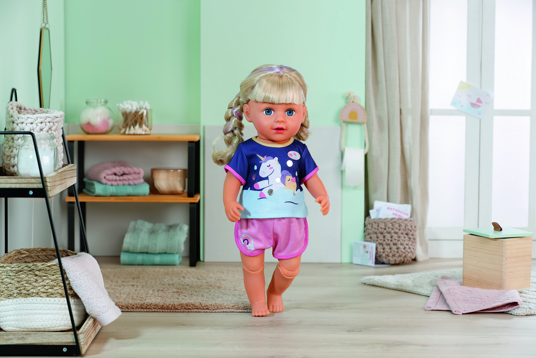 Lalka w koszulce z jednorożcem i różowych spodenkach stoi w łazience. Tła: półki, ręczniki i okno.