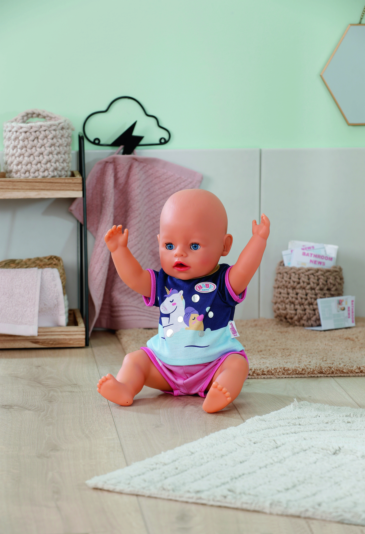 Lalka siedzi z uniesionymi ramionami. Ma na sobie koszulę i szorty. Za nią drewniana półka i dekoracja chmury. Tło jest jasne.