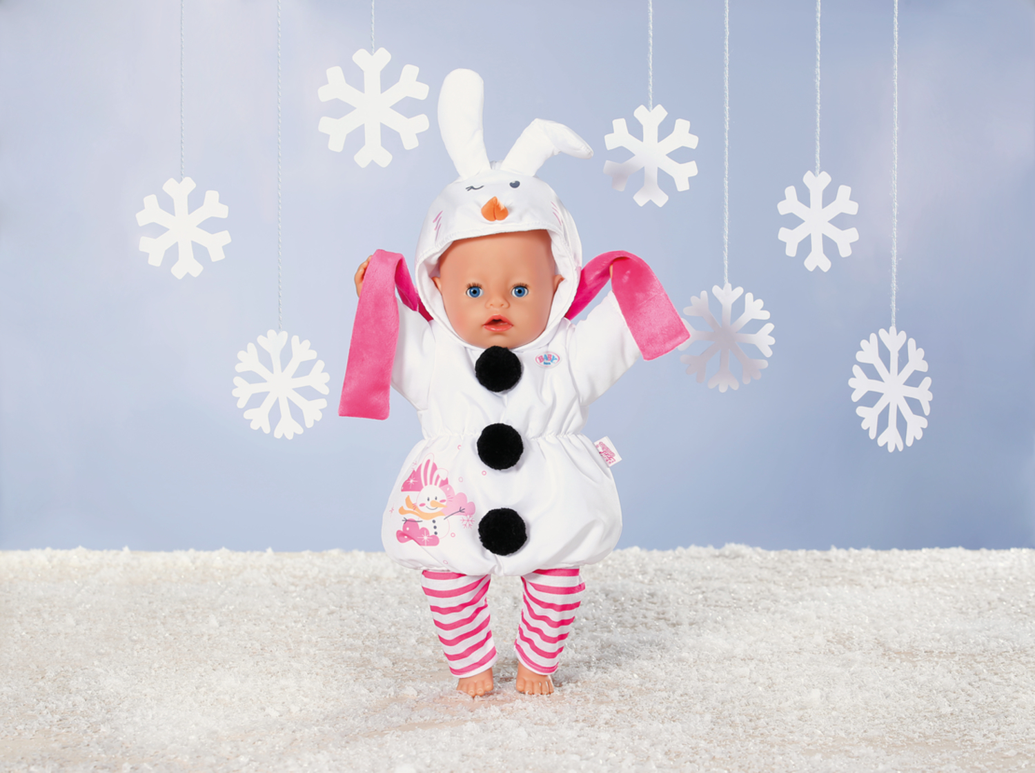Lalka w kostiumie bałwana z białym strojem, czarnymi guzikami, różowym szalikiem i paskami. Płatki śniegu i śnieżne tło.