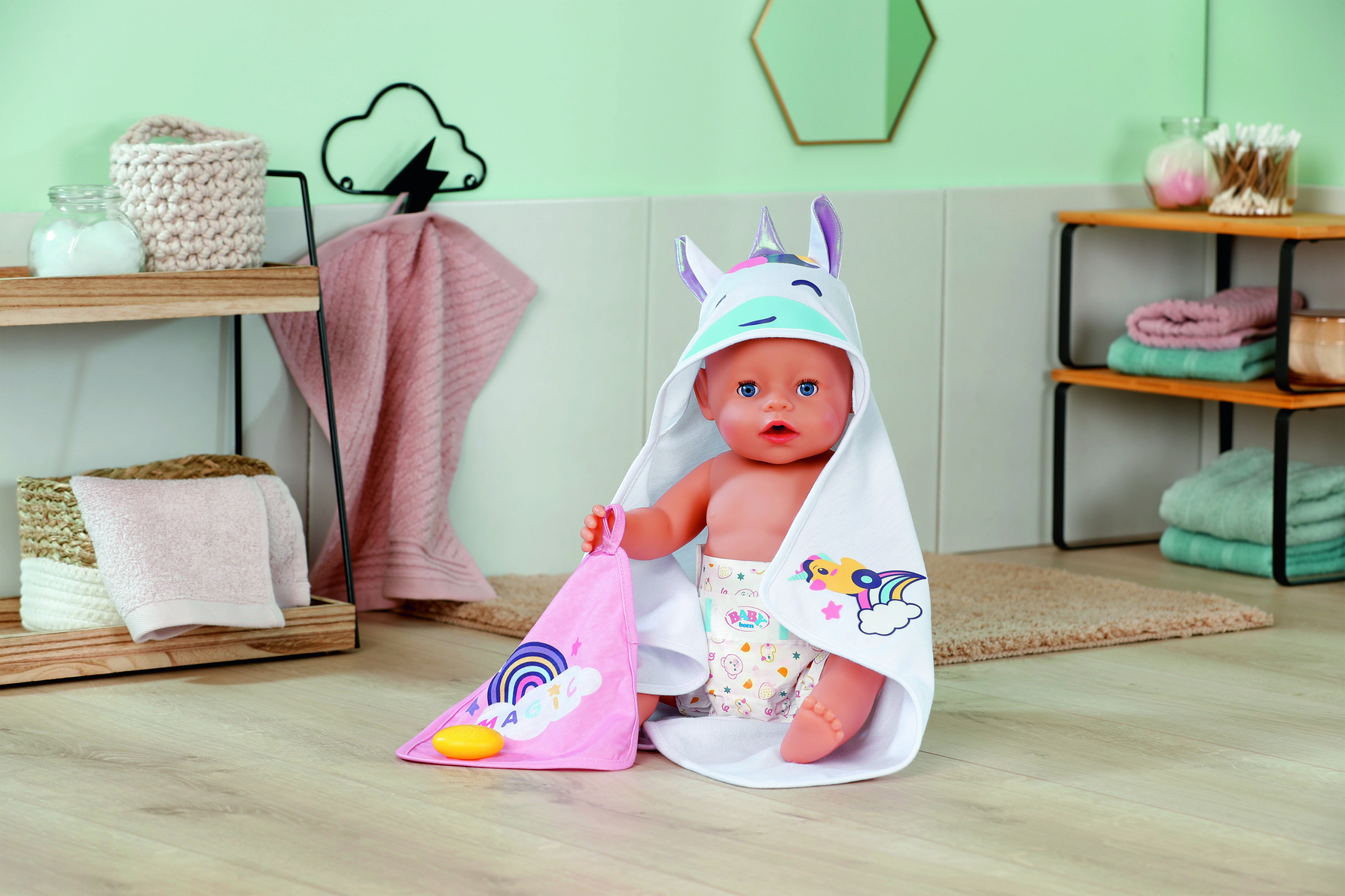 Lalka w ręczniku z motywem jednorożca, trzymająca różowy ręcznik, w łazience z półkami i ręcznikami.