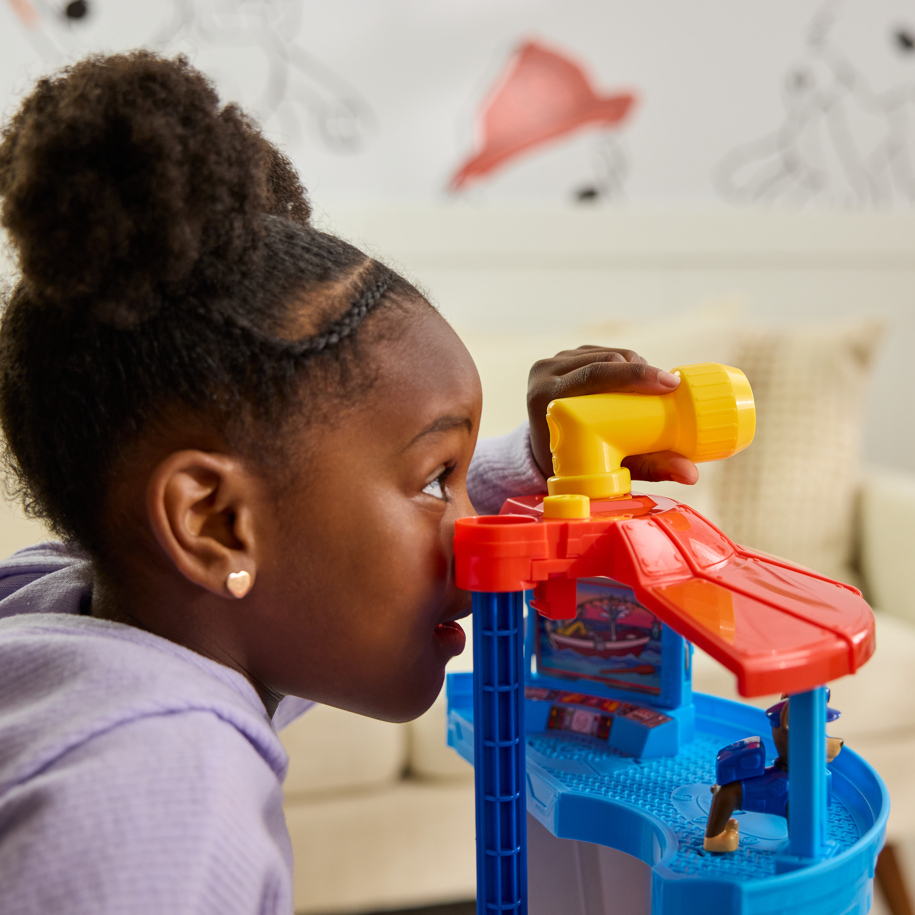 Młoda dziewczyna patrzy przez żółty teleskop na niebiesko-czerwonej zabawce. Ma na sobie fioletową koszulę i włosy spięte w kok.