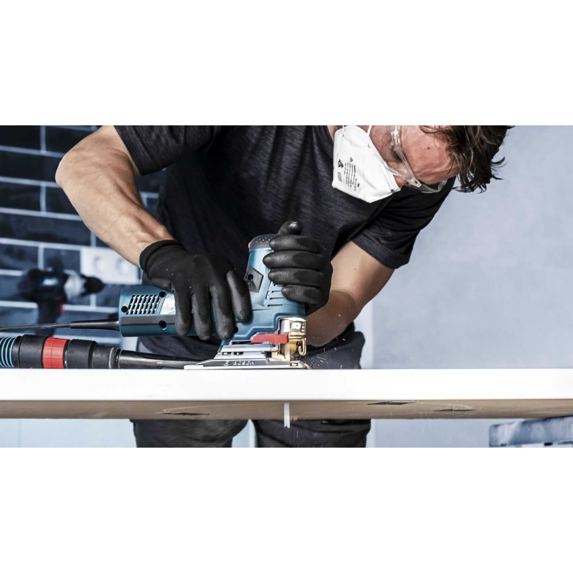 Osoba w masce i rękawicach używa wyrzynarki do cięcia białej płyty. Widoczne są iskry. Tło to ceglana ściana.