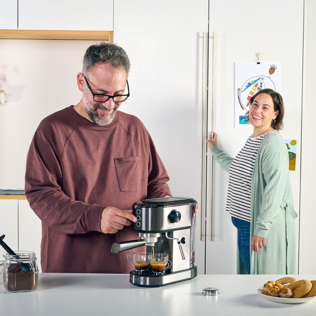 Mężczyzna robi kawę ekspresem, a kobieta się uśmiecha. Na blacie filiżanki, ciastka i zmielona kawa.