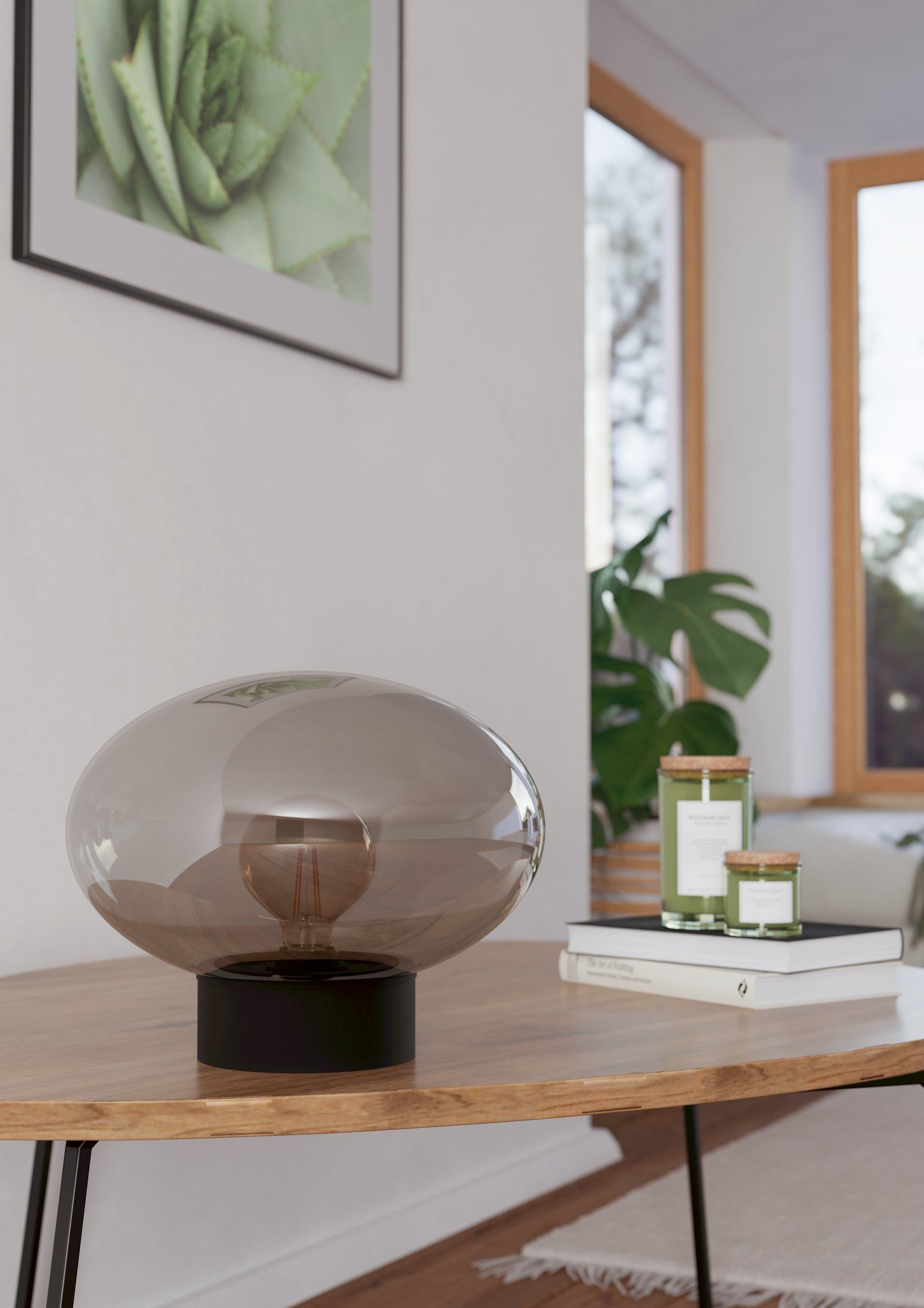 Eine ovale Lampe steht auf einem Holztisch mit Kerzen und Büchern. Ein gerahmter Sukkulenten-Druck hängt an der Wand.
