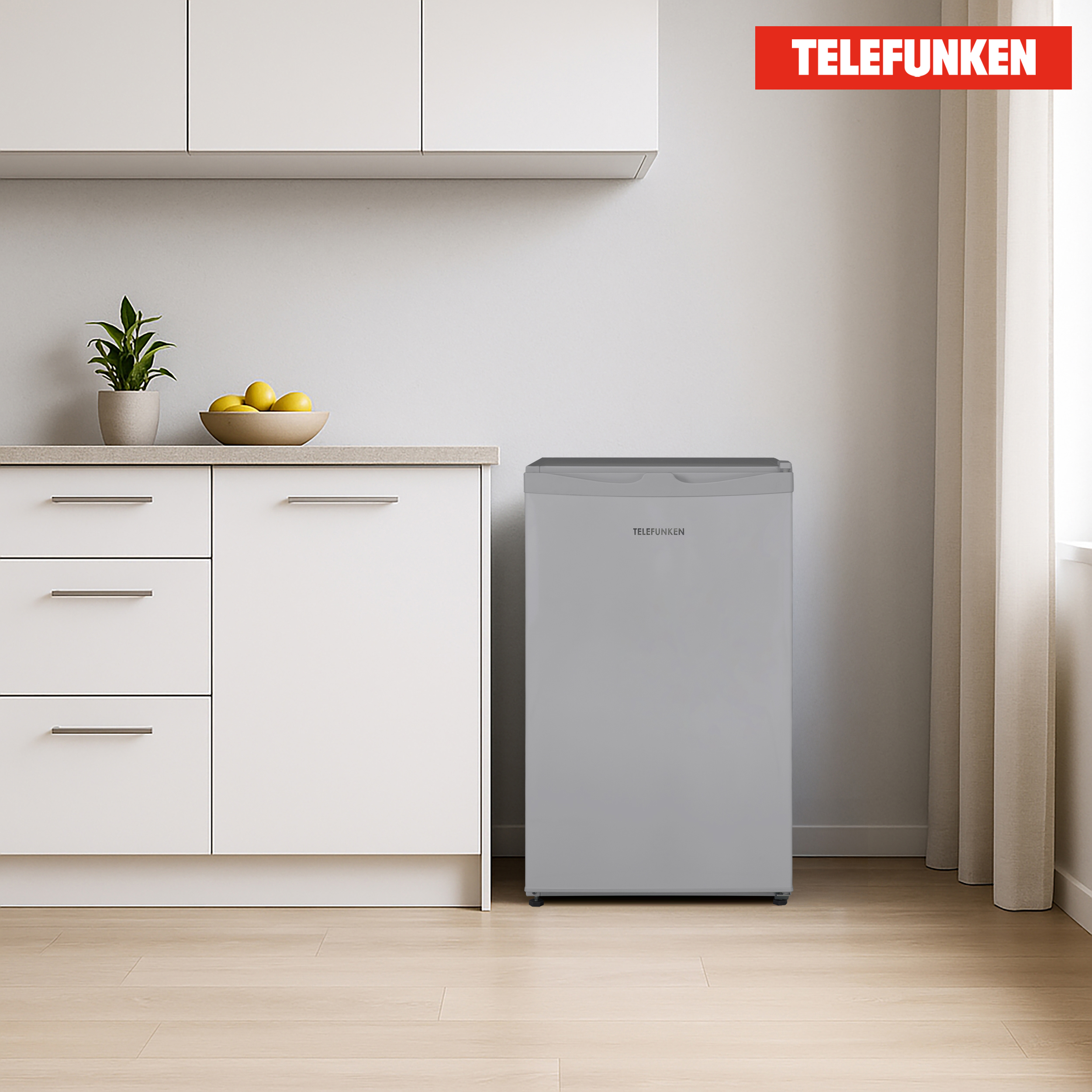 Ein grauer Minikühlschrank in einer Küche, Telefunken-Logo darauf. Weiße Schränke und gelbe Zitronen in einer Schüssel sind ebenfalls sichtbar.