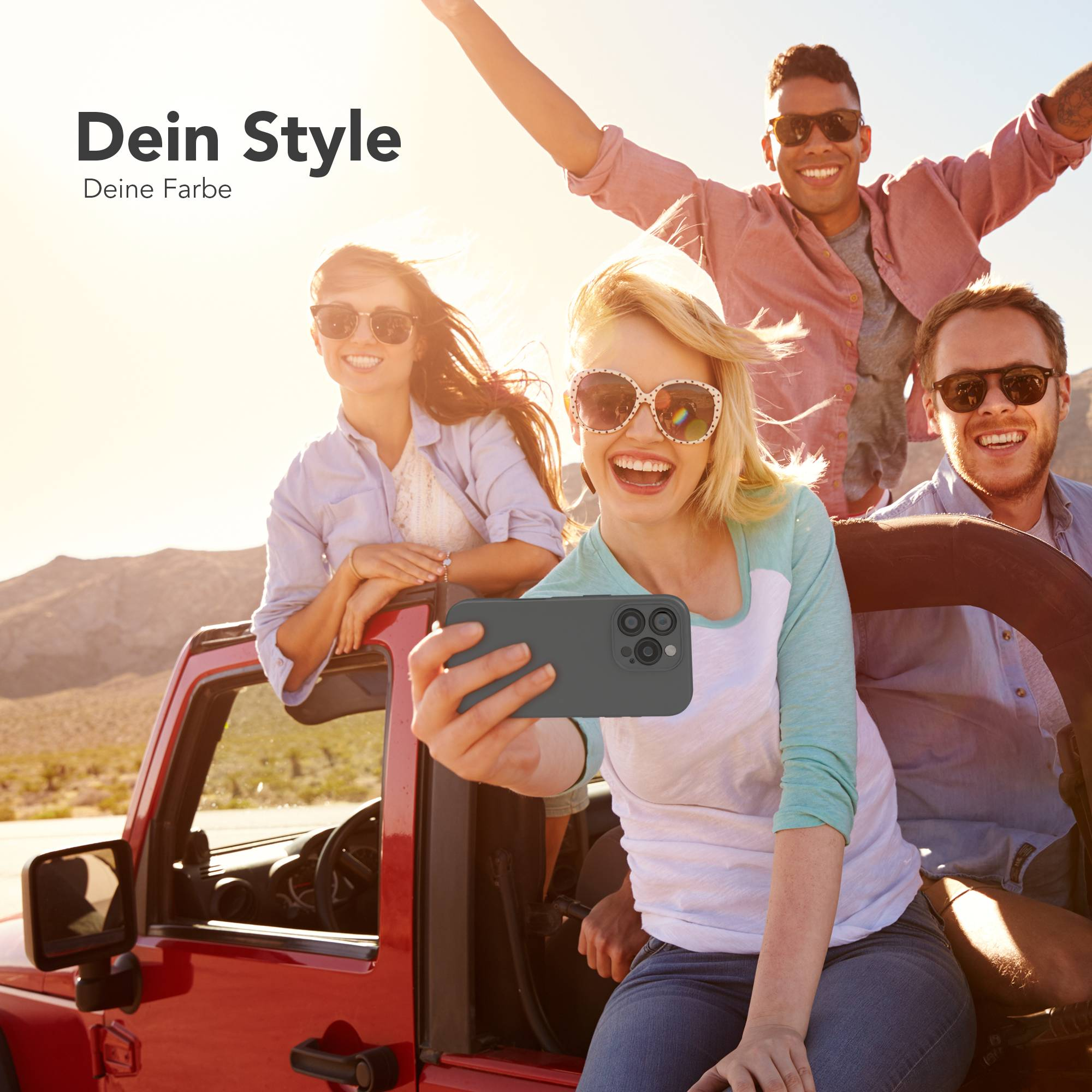 Vier junge Erwachsene in einem roten Jeep machen ein Selfie. Sie tragen Sonnenbrillen, lächeln, mit Berg im Hintergrund.