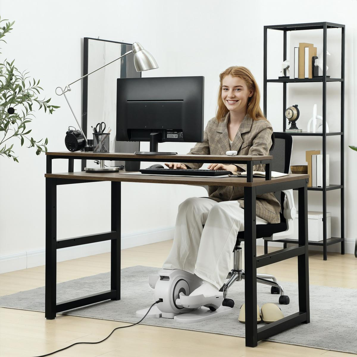Frau am Schreibtisch, benutzt einen Computer. Sie trägt eine Jacke und weiße Hose. Ein Heimtrainer steht unter dem Tisch. Hintergrund mit Regal.