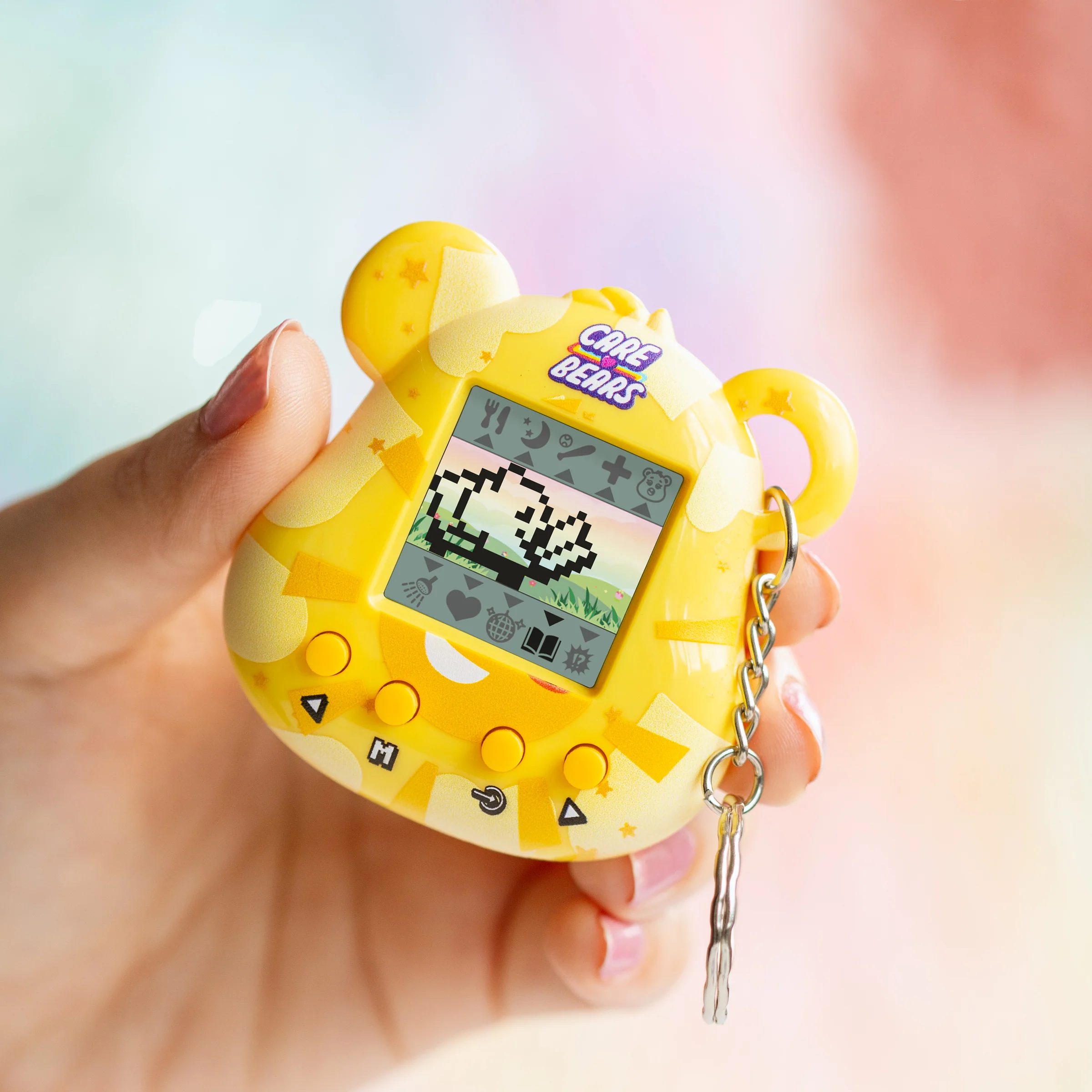 Een gele Care Bears-speelgoed wordt door een hand vastgehouden. Het scherm toont een gepixeleerde beer. Een zilveren ketting is bevestigd.