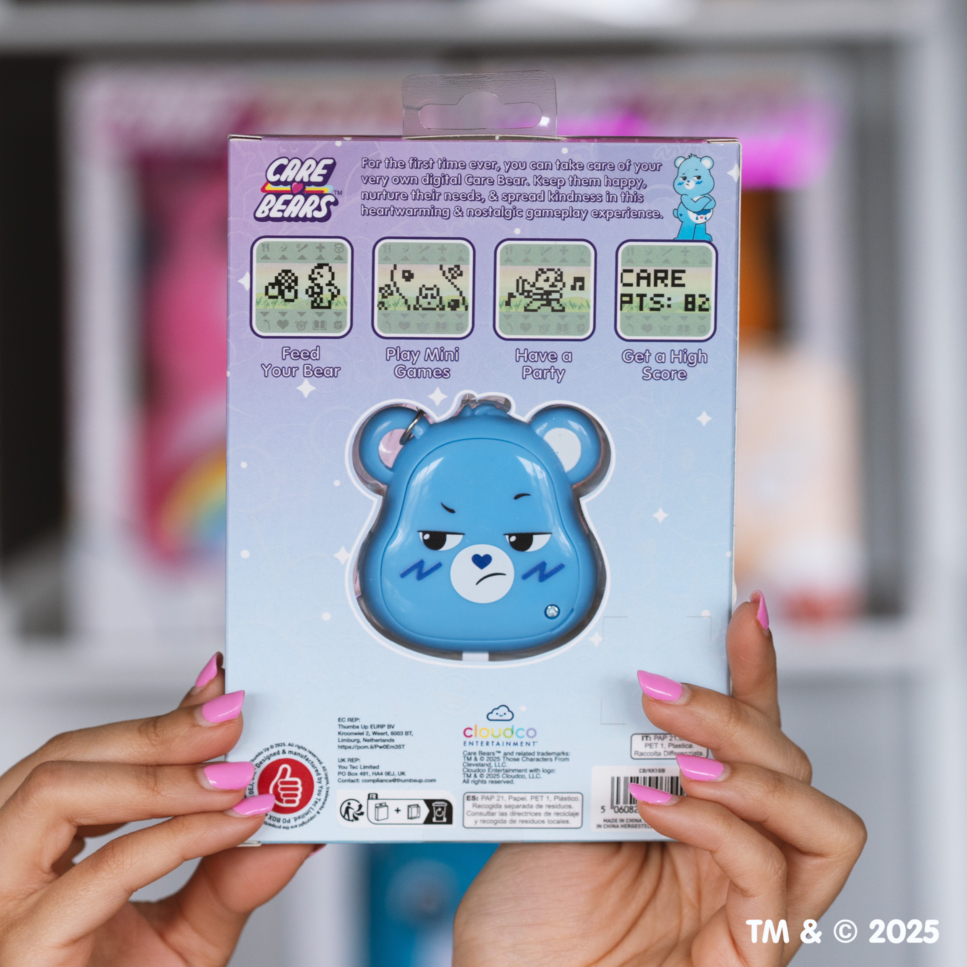 Een persoon houdt een digitaal Care Bears-speeltje vast. De verpakking is lichtblauw met een Care Bear-afbeelding. Roze nagels zijn zichtbaar.