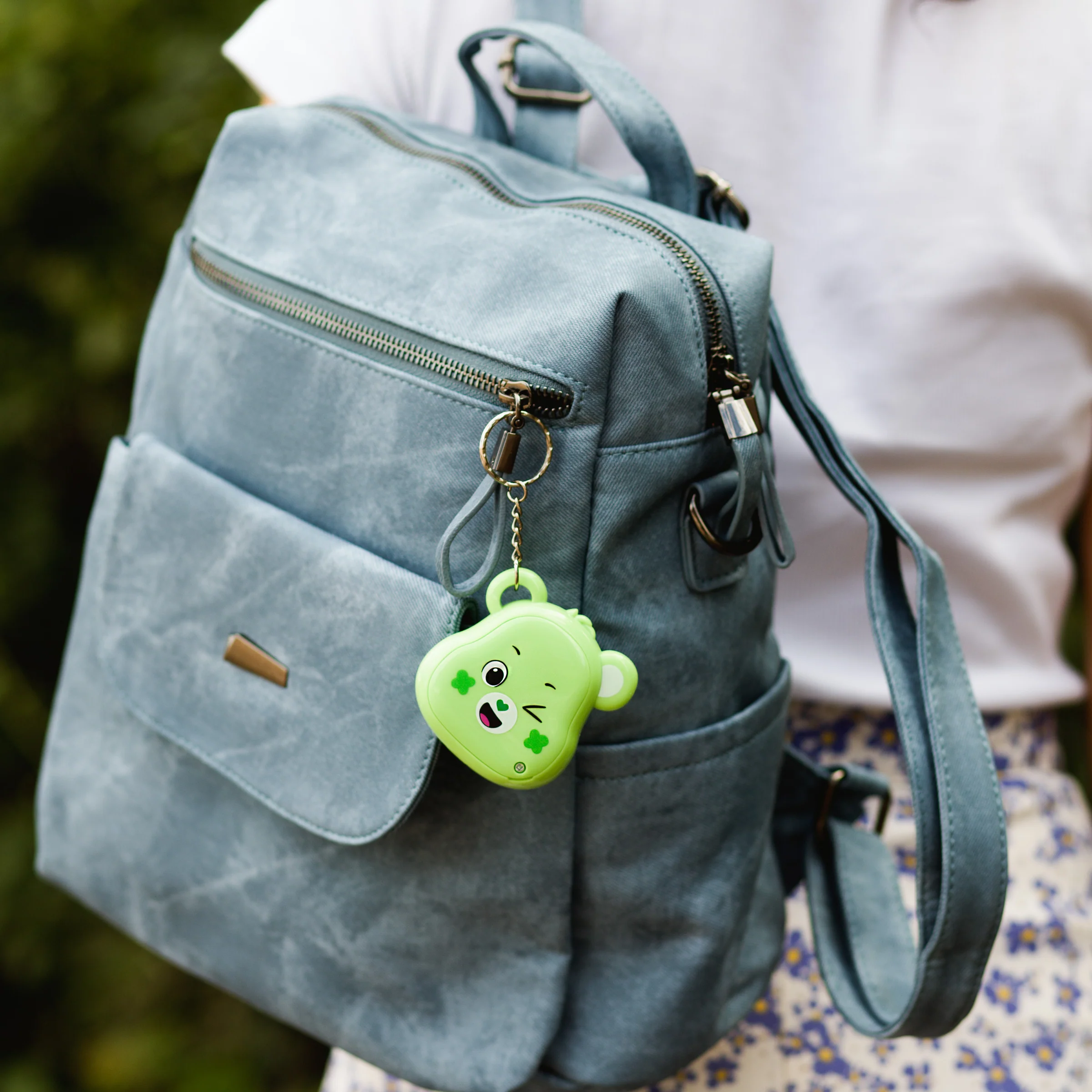 Een blauwe rugzak met een groene beren sleutelhanger. De achtergrond toont een wit shirt en een bloemenrok.