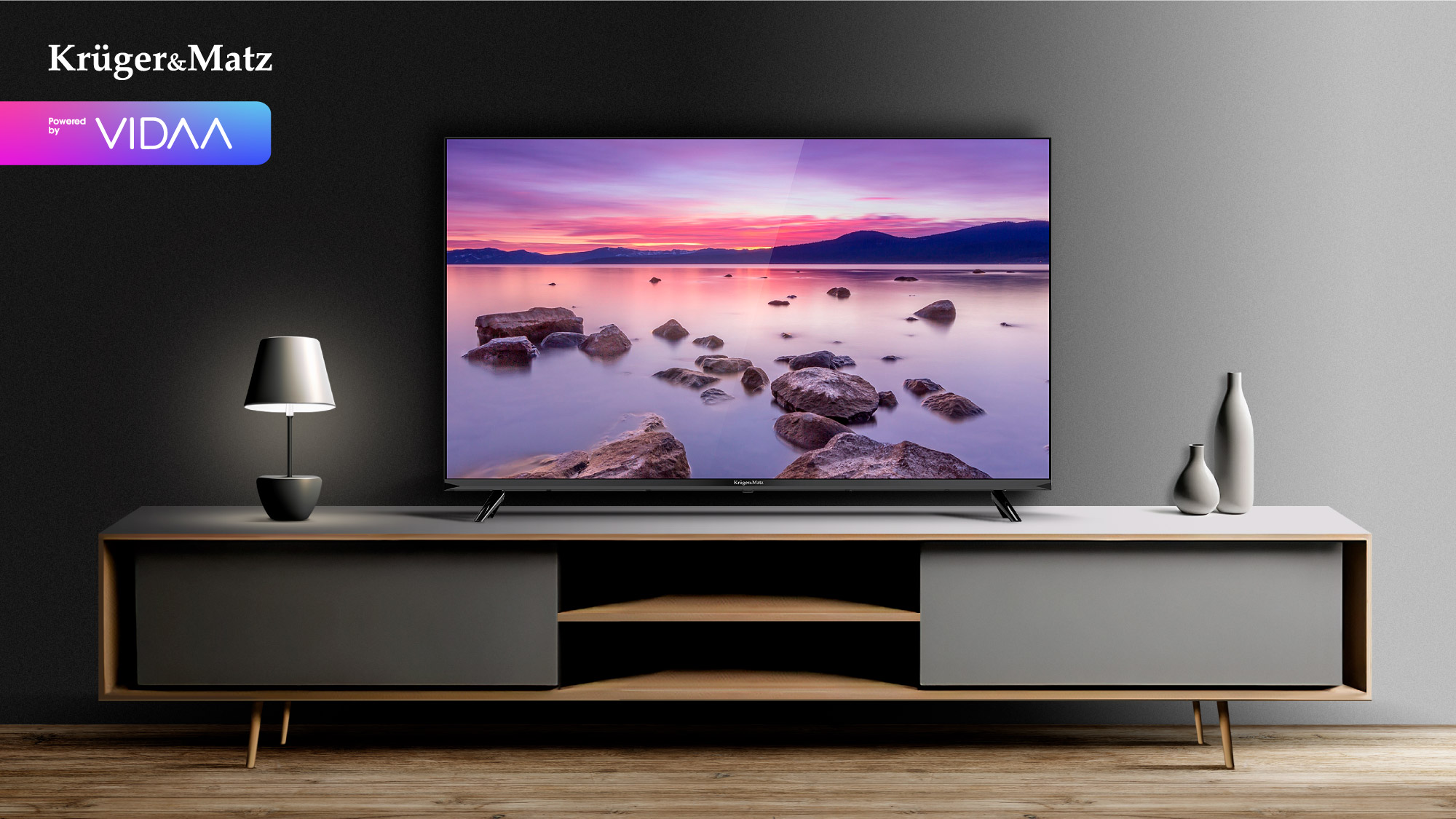 Telewizor wyświetla zachód słońca nad wodą i skałami. Stoi na szarej szafce. Na szafce znajdują się również lampa i wazony.